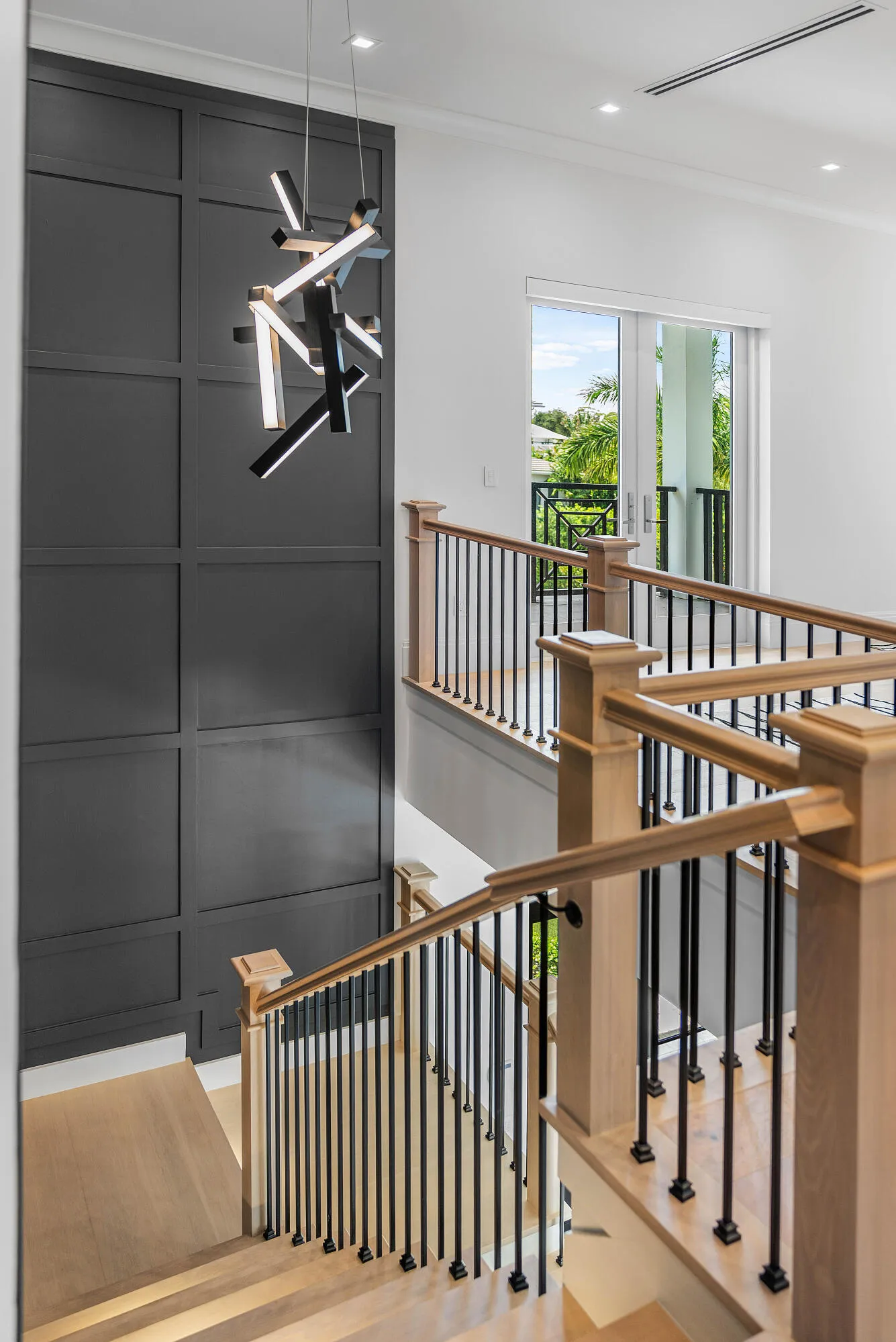 Modern chandelier illuminating a paneled accent wall, light wood-finish stairs, and black metal balusters with wood handrails
