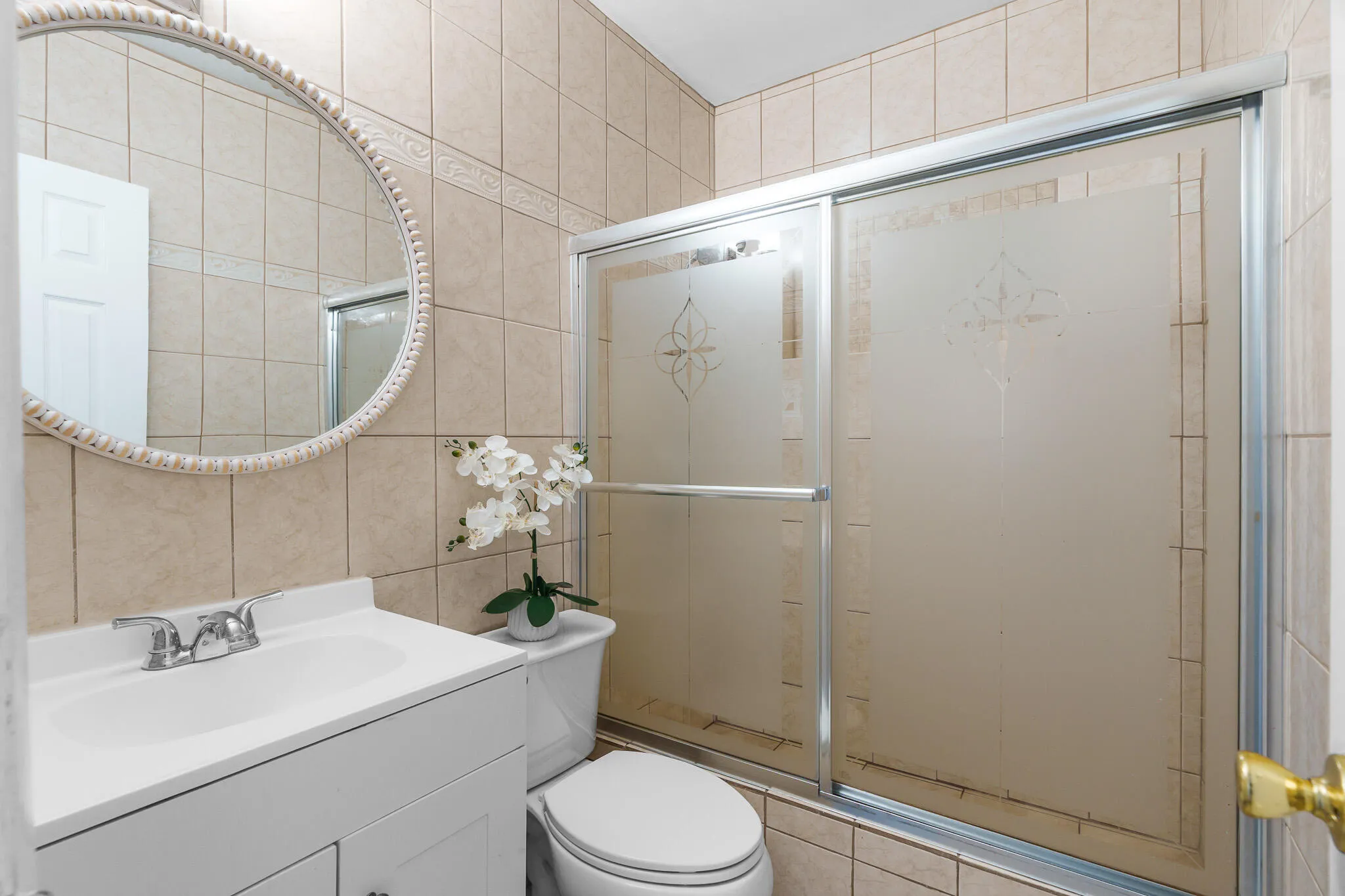 Tiled bathroom with a white vanity featuring a single basin sink and chrome faucet, an oval mirror with a decorative frame, and a shower enclosure with frosted glass panels