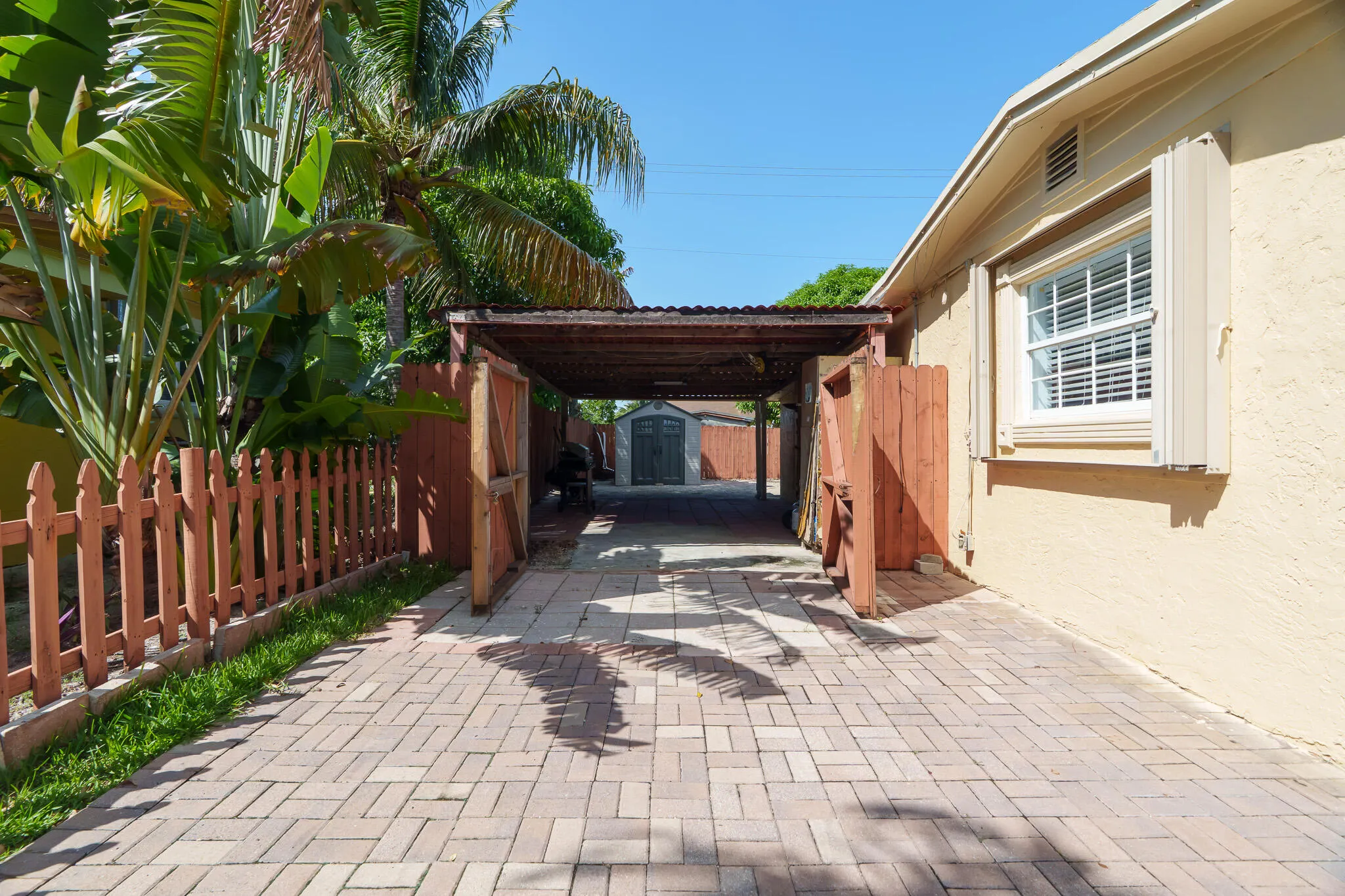 Paver driveway leading to a covered carport, featuring a picket fence and tropical landscaping