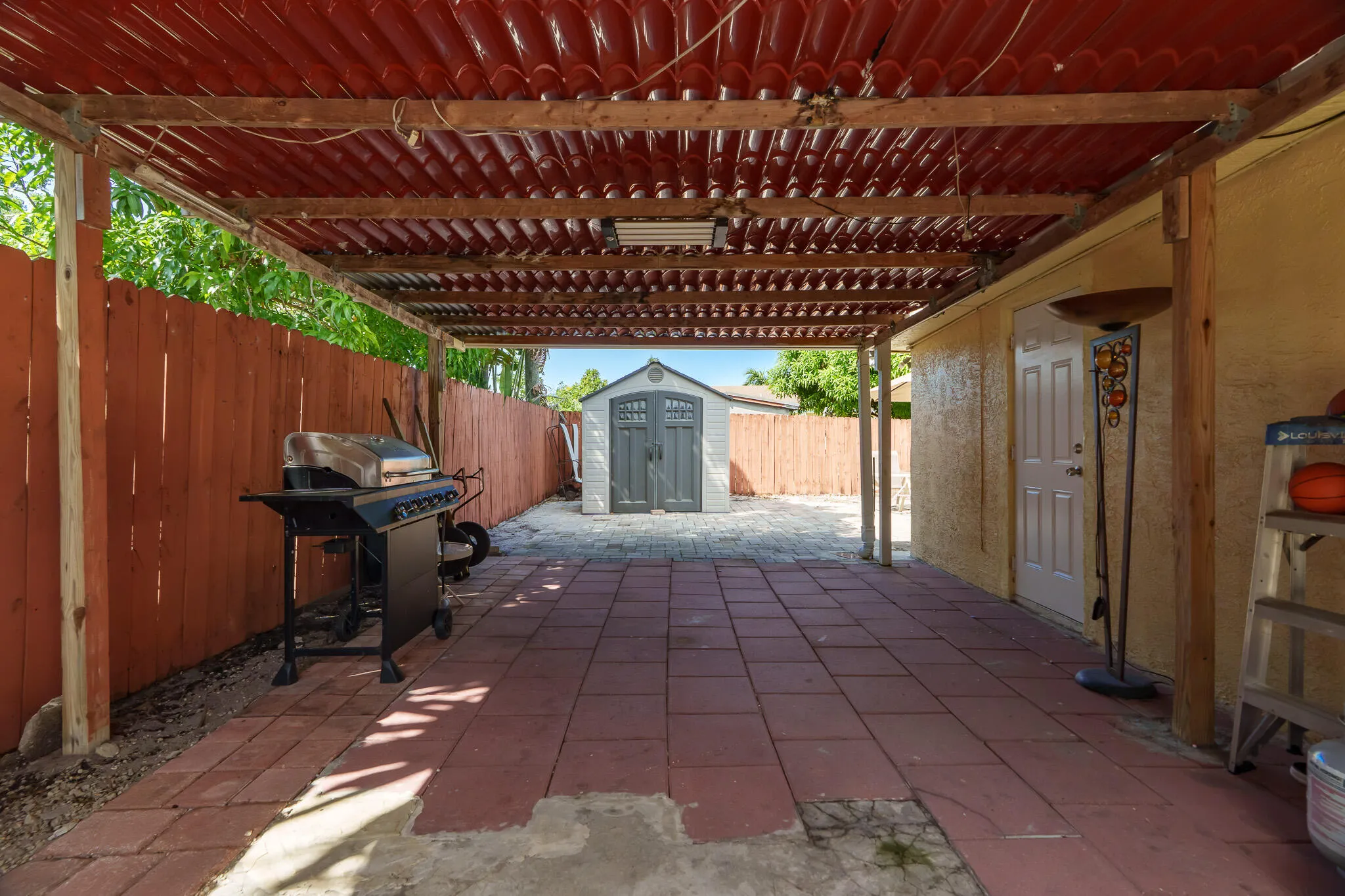 Covered carport area with a red corrugated roof, wood-beam supports, and red paver flooring