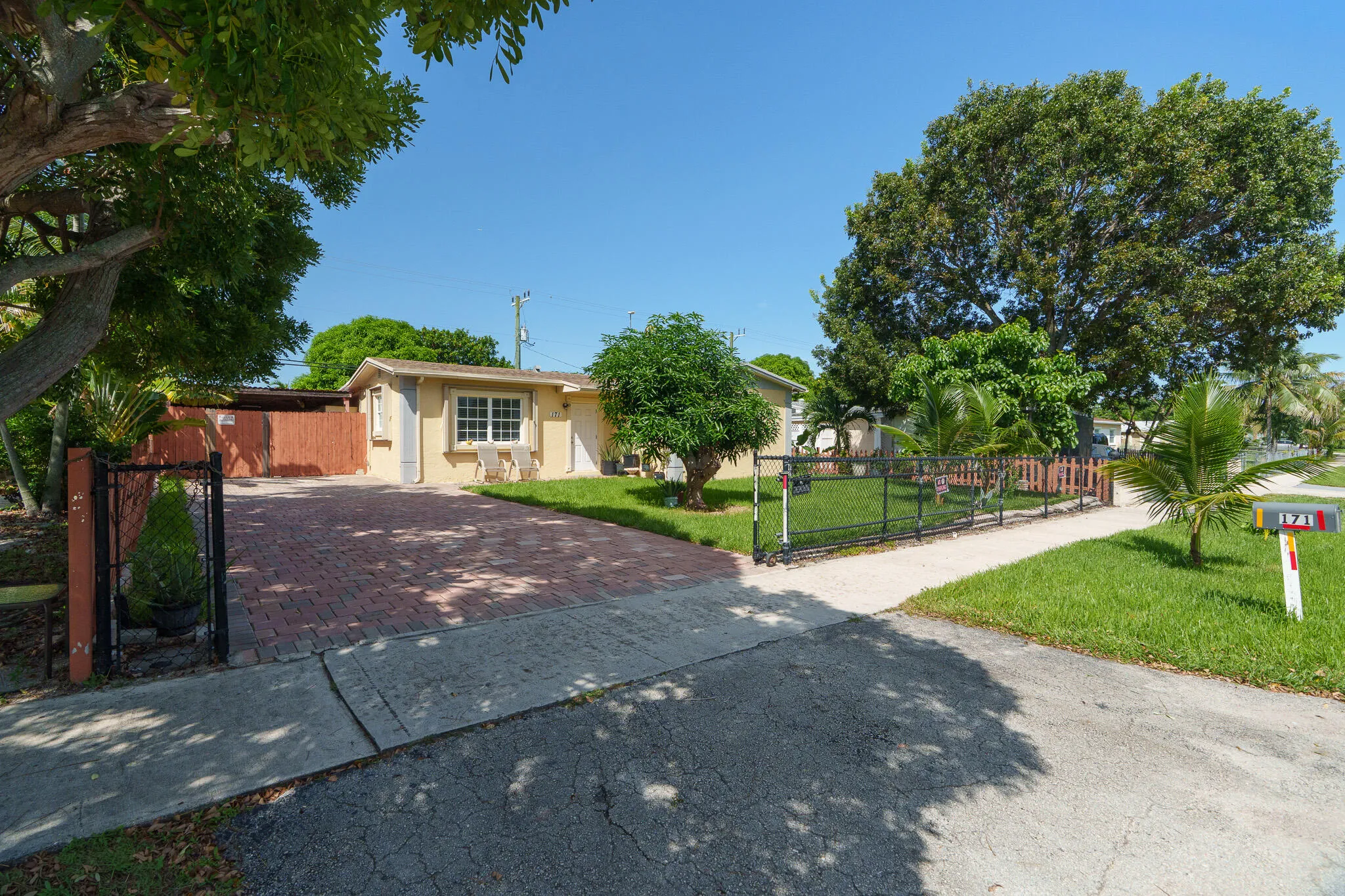 Paved driveway, mature trees, green lawn, fenced front yard, and light-colored exterior