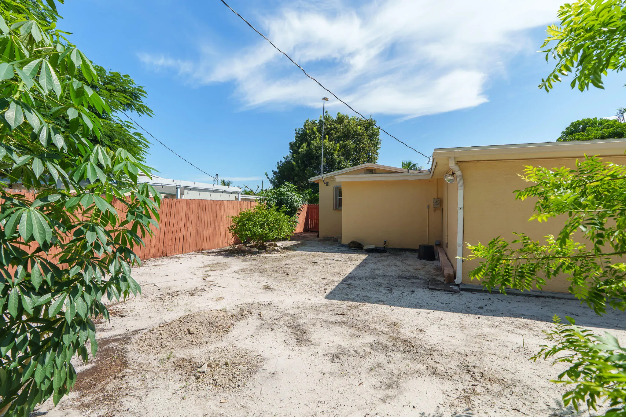 Expansive outdoor space featuring a partially paved yard, mature perimeter foliage, and a covered side area