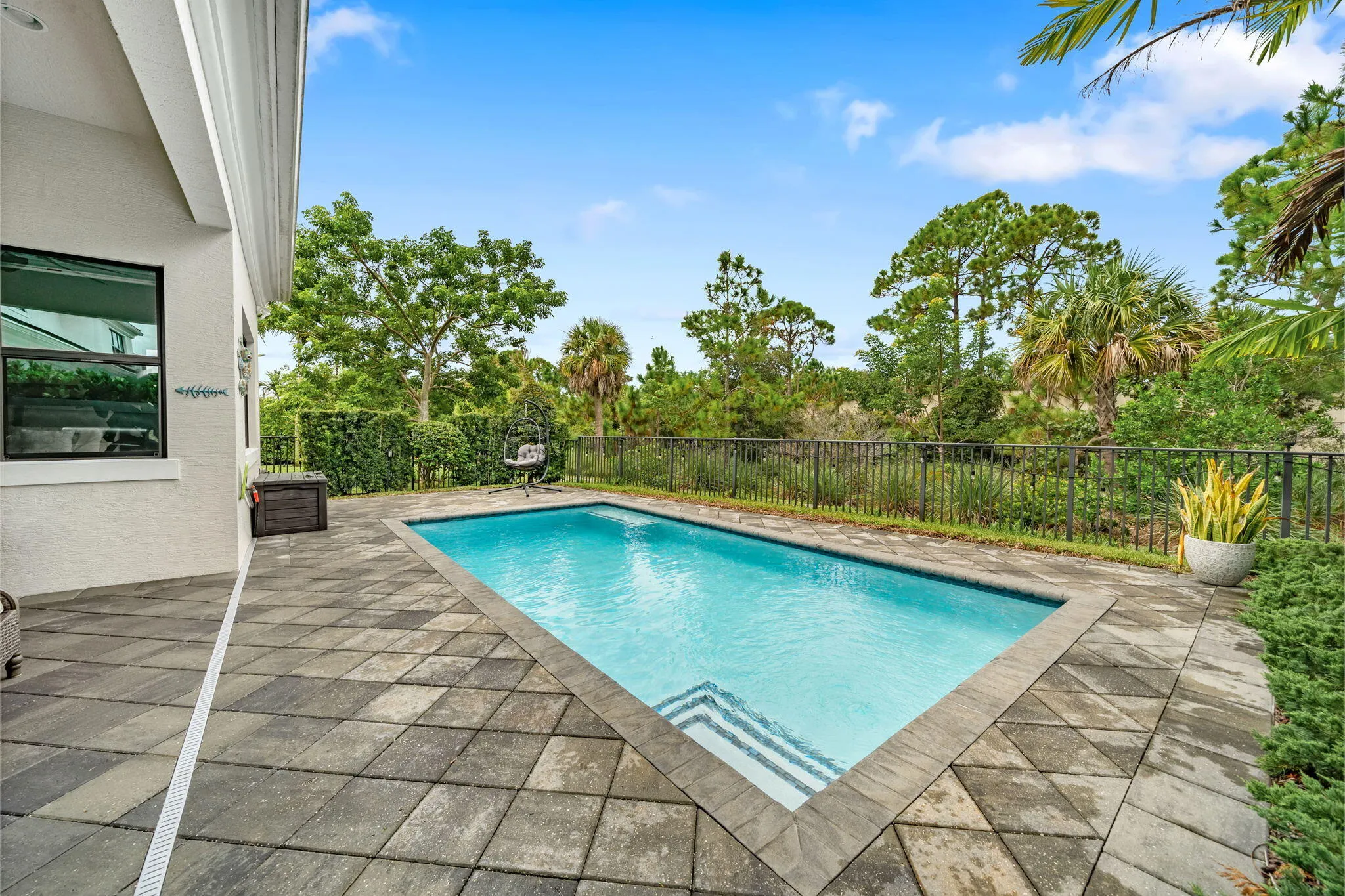 Private swimming pool with integrated steps, surrounded by a paver patio and a black metal fence, with dense perimeter landscaping and preserve