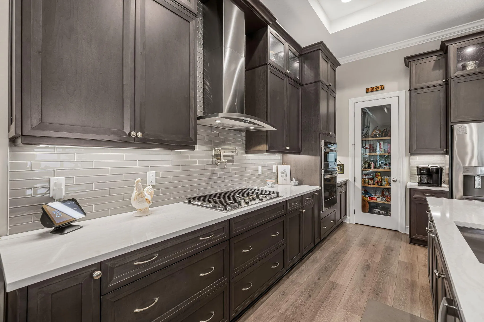 Dark wood cabinetry, stainless steel range hood, built-in gas cooktop, glass tile backsplash, under cabinet lighting, recessed lighting, and a pantry with a glass-paneled door
