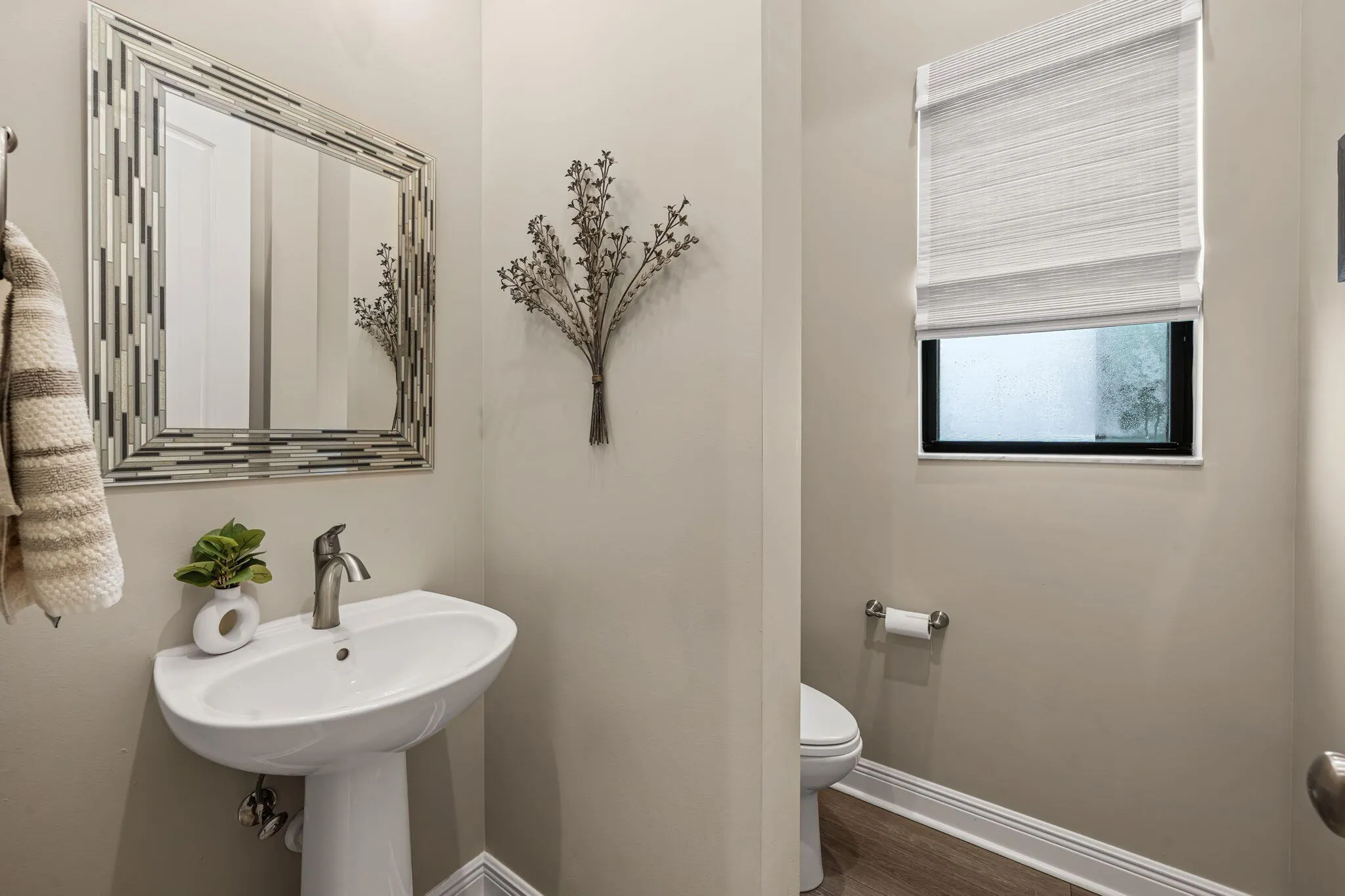 Pedestal sink with a brushed nickel faucet, framed mirror with a mosaic border, and a window with a textured Roman shade