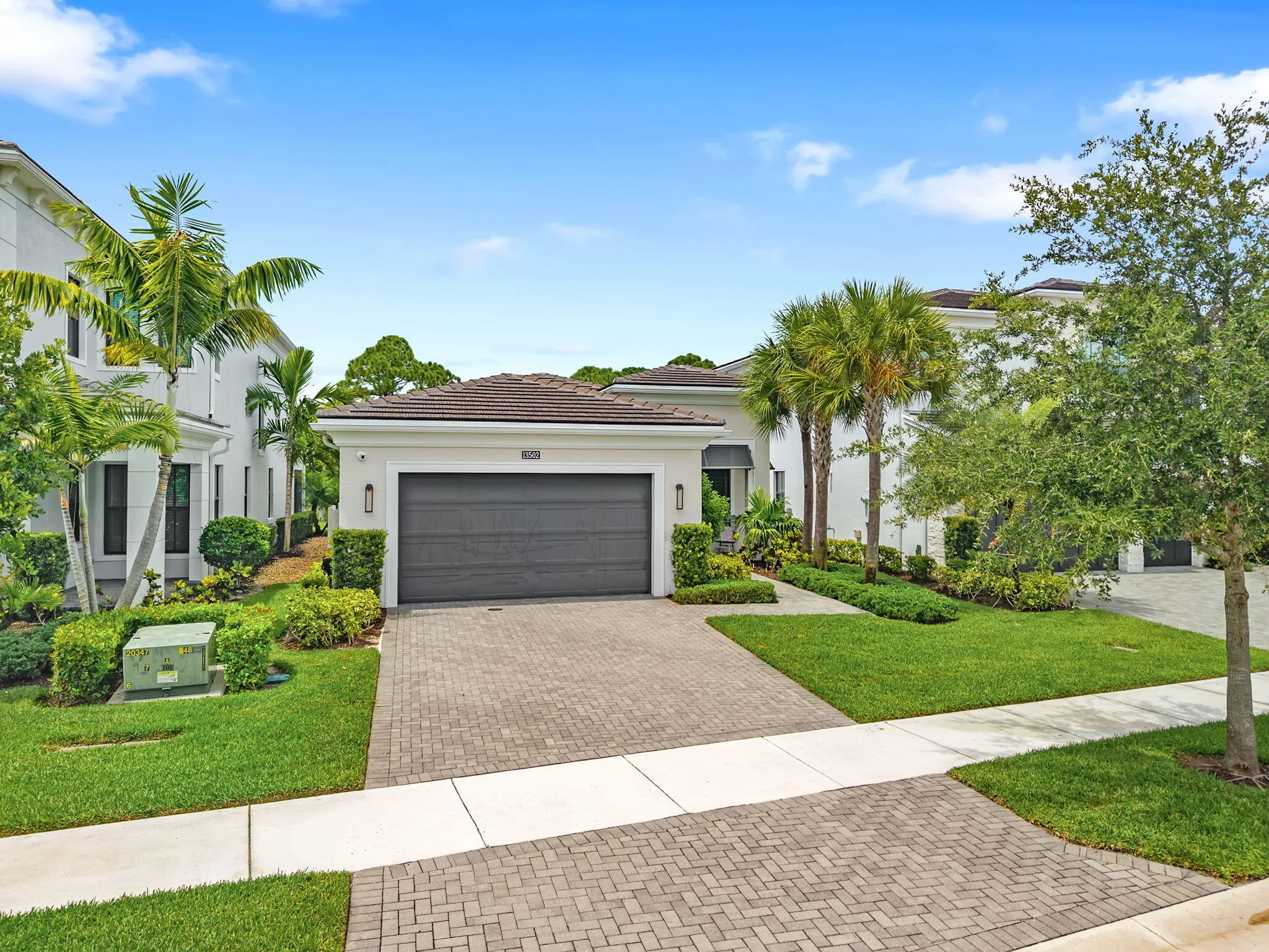 White stucco exterior with a dark tile roof, paver driveway, and manicured landscaping featuring mature trees