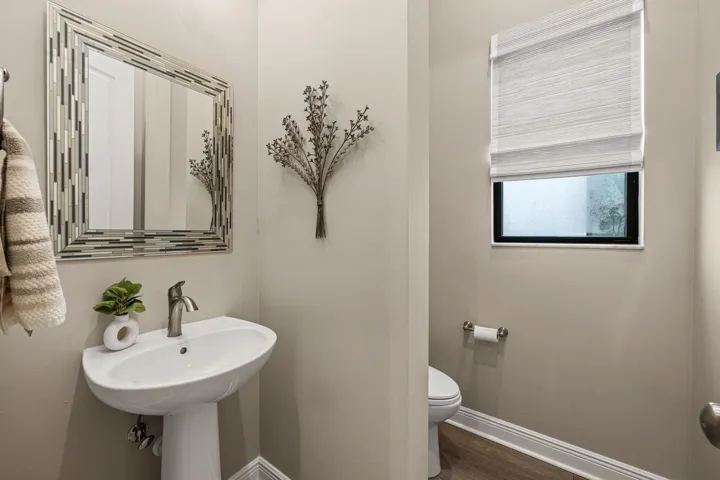 Pedestal sink with a brushed nickel faucet, framed mirror with a mosaic border, and a window with a textured Roman shade