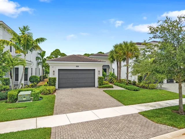 White stucco exterior with a dark tile roof, paver driveway, and manicured landscaping featuring mature trees