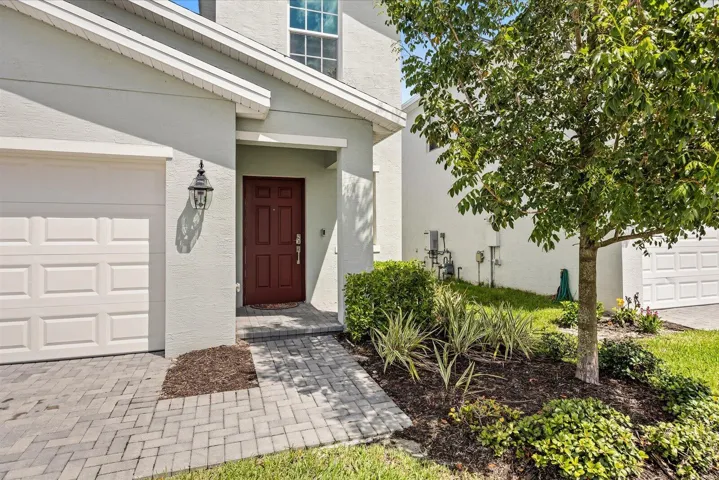 Light-toned stucco exterior with a paneled garage door, a patterned paver walkway, and mature landscaping