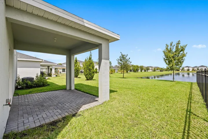 Covered patio with paver flooring, expansive lawn, and black fence bordering a water feature