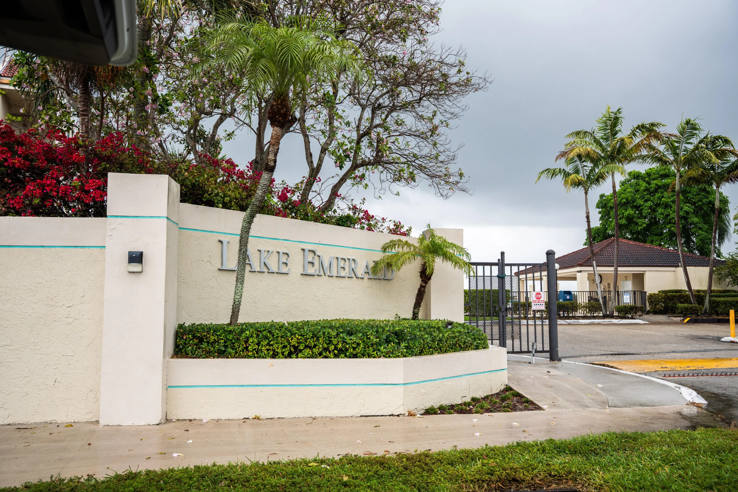 Community entrance featuring a privacy gate, stucco entry wall with recessed lettering, mature palm trees, and lush landscaping
