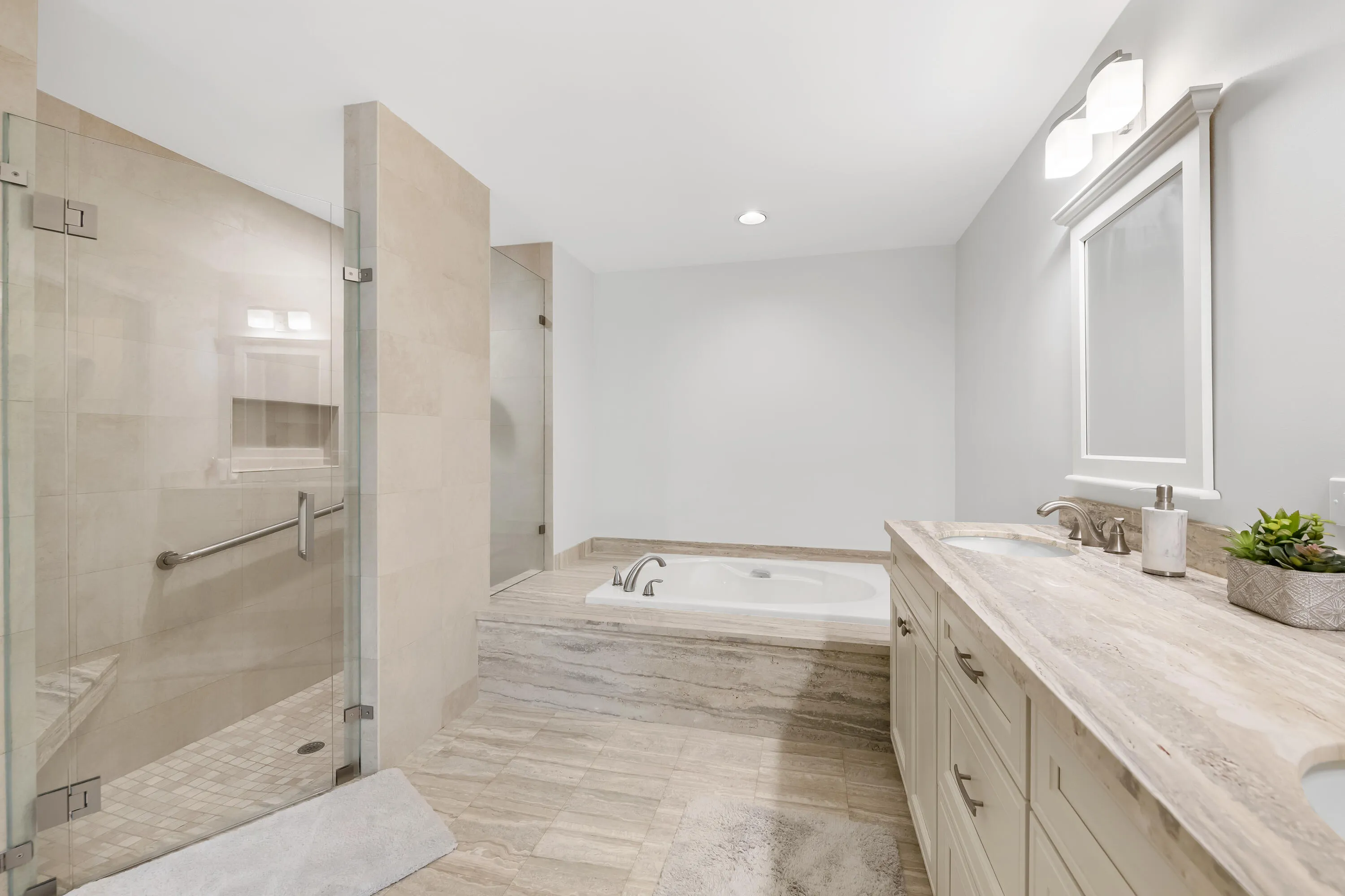 Spacious bathroom featuring a dual vanity with stone countertops, a built-in soaking tub, and a glass-enclosed shower with a bench and grab bar