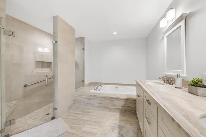 Spacious bathroom featuring a dual vanity with stone countertops, a built-in soaking tub, and a glass-enclosed shower with a bench and grab bar