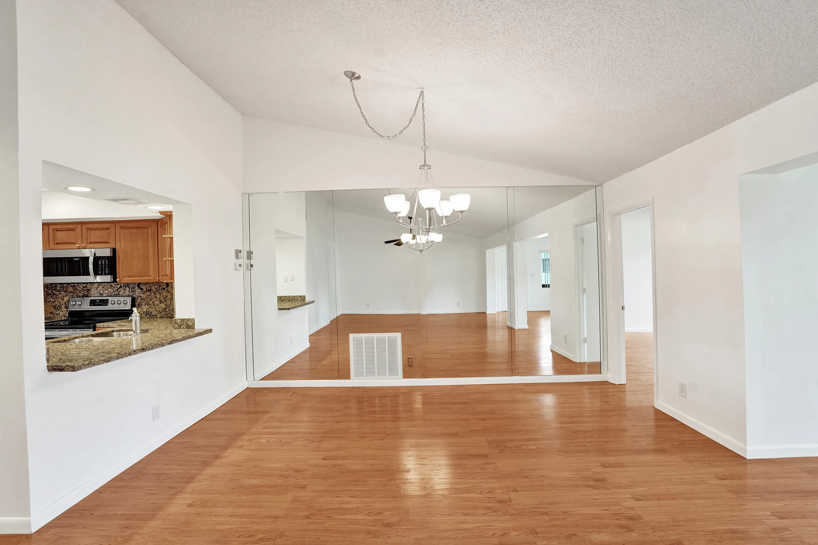Vaulted ceiling with a chandelier, wood-finish flooring, kitchen pass-through, and full-wall mirrors