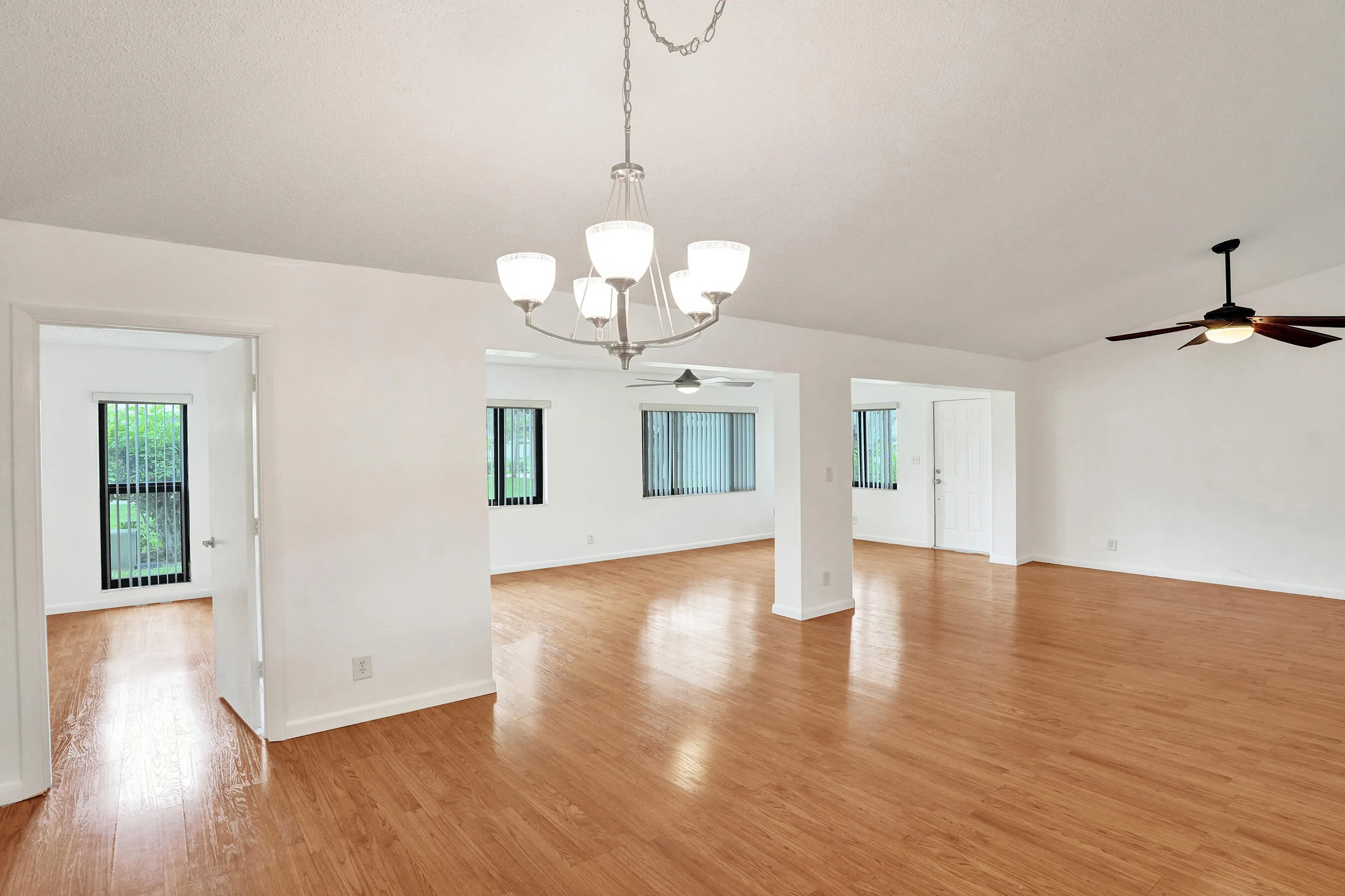 Wood-finish flooring throughout, bright white walls, multiple windows with vertical blinds, and a ceiling fan with integrated lighting