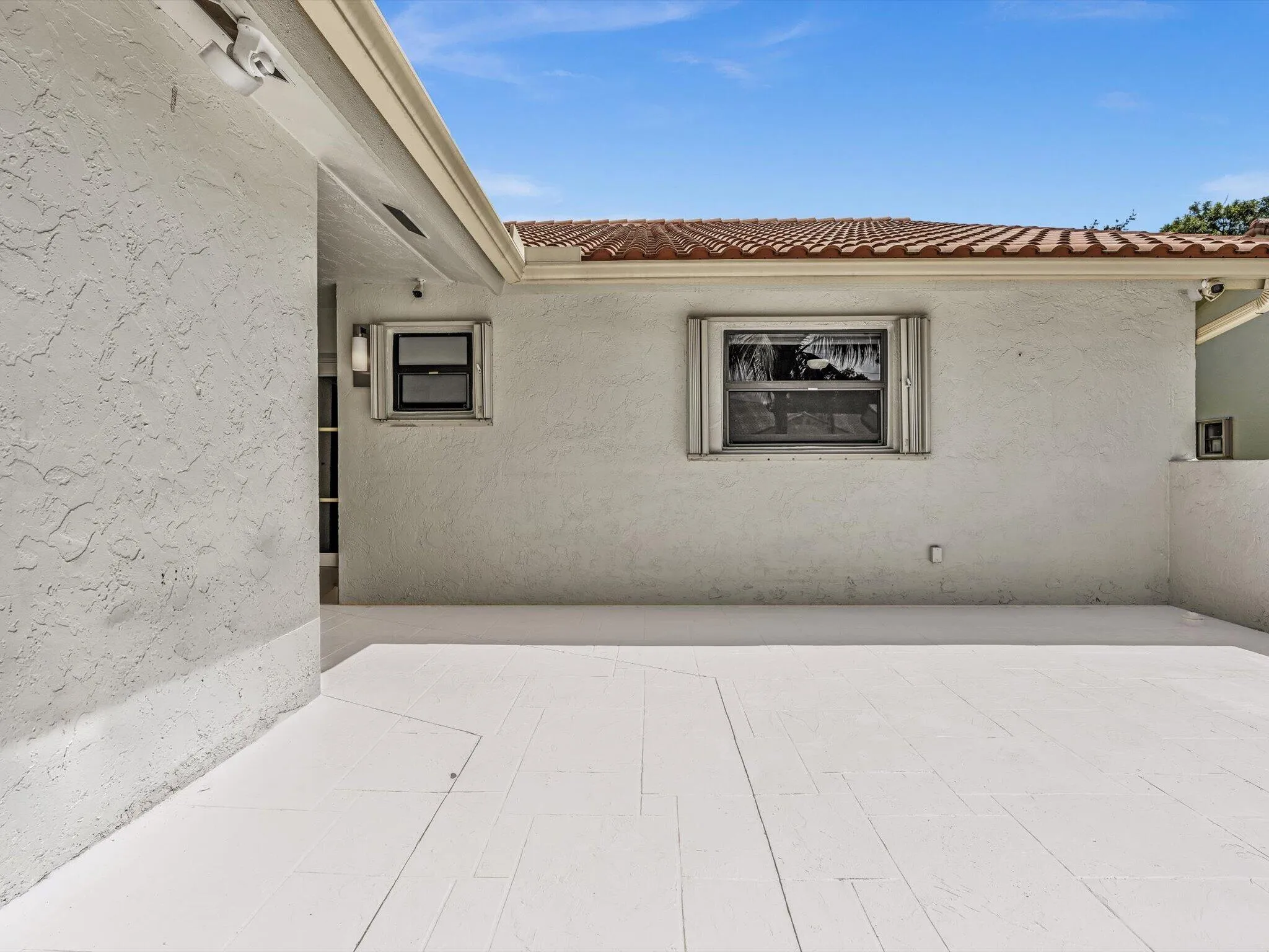 Exterior stucco finish, tile roof, window with louvered shutters, outdoor wall light, and an expansive concrete patio