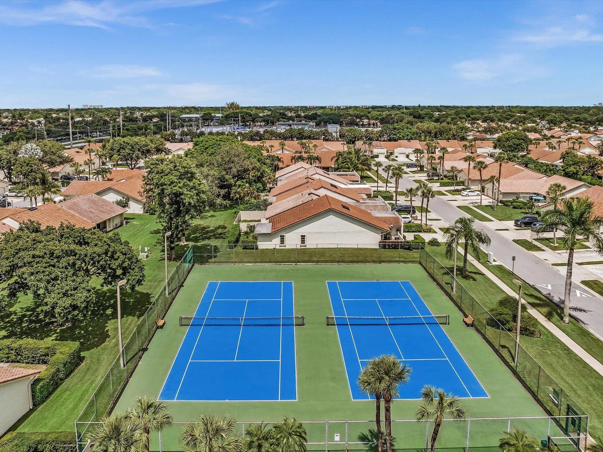 Two hard tennis courts with blue playing surfaces and green surrounds, surrounded by fencing