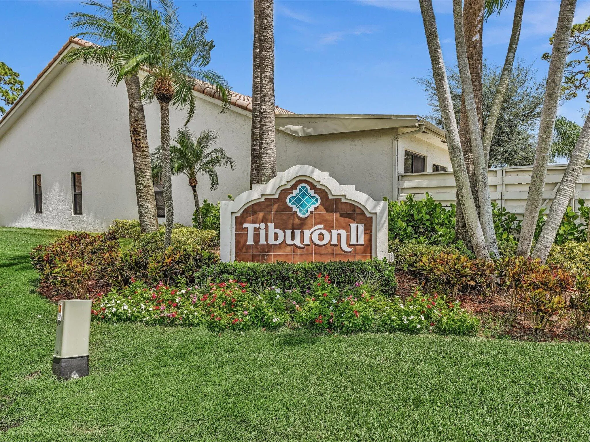 Community entrance sign featuring decorative tile accents, lush landscaping with flowering plants, mature palm trees, and manicured lawn areas