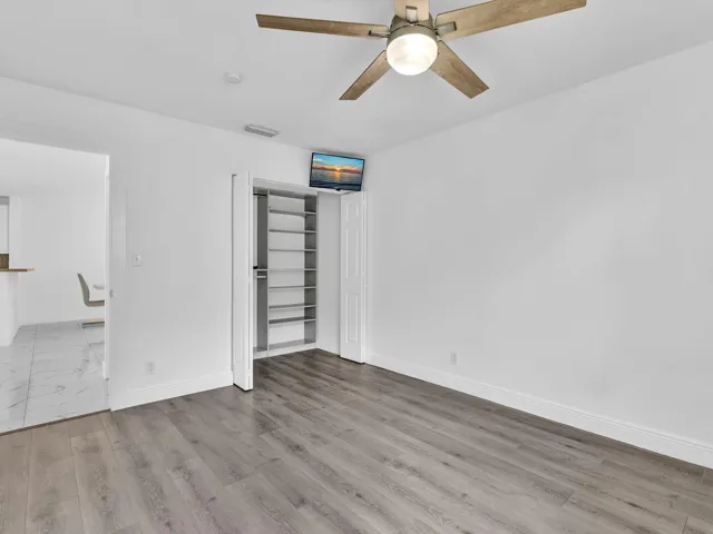 Wood-finish flooring, ceiling fan with integrated lighting, built-in closet shelving, and a clean white wall finish