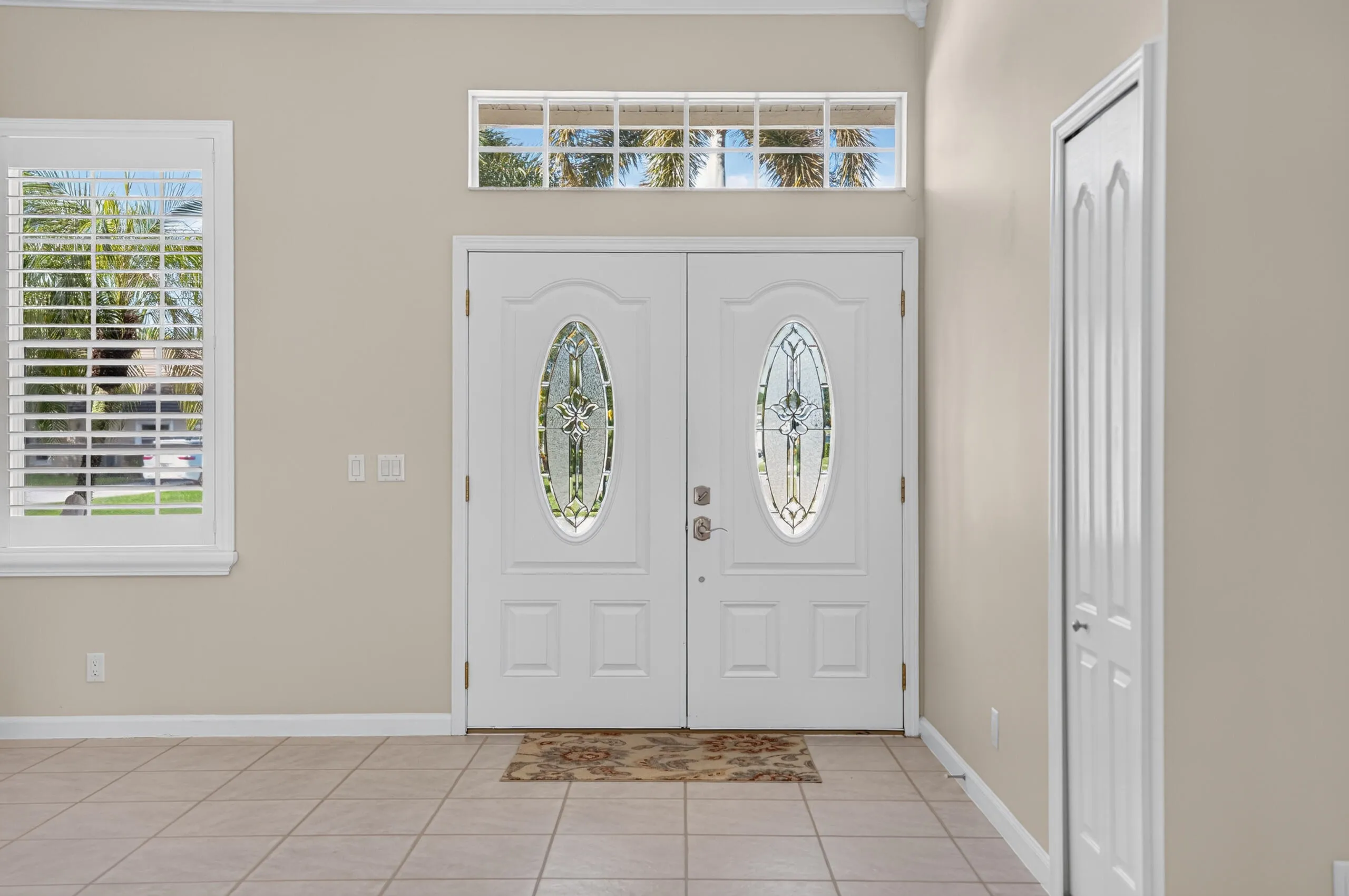 Double entry doors with decorative oval glass inserts, transom window, window with interior shutters, and tile flooring