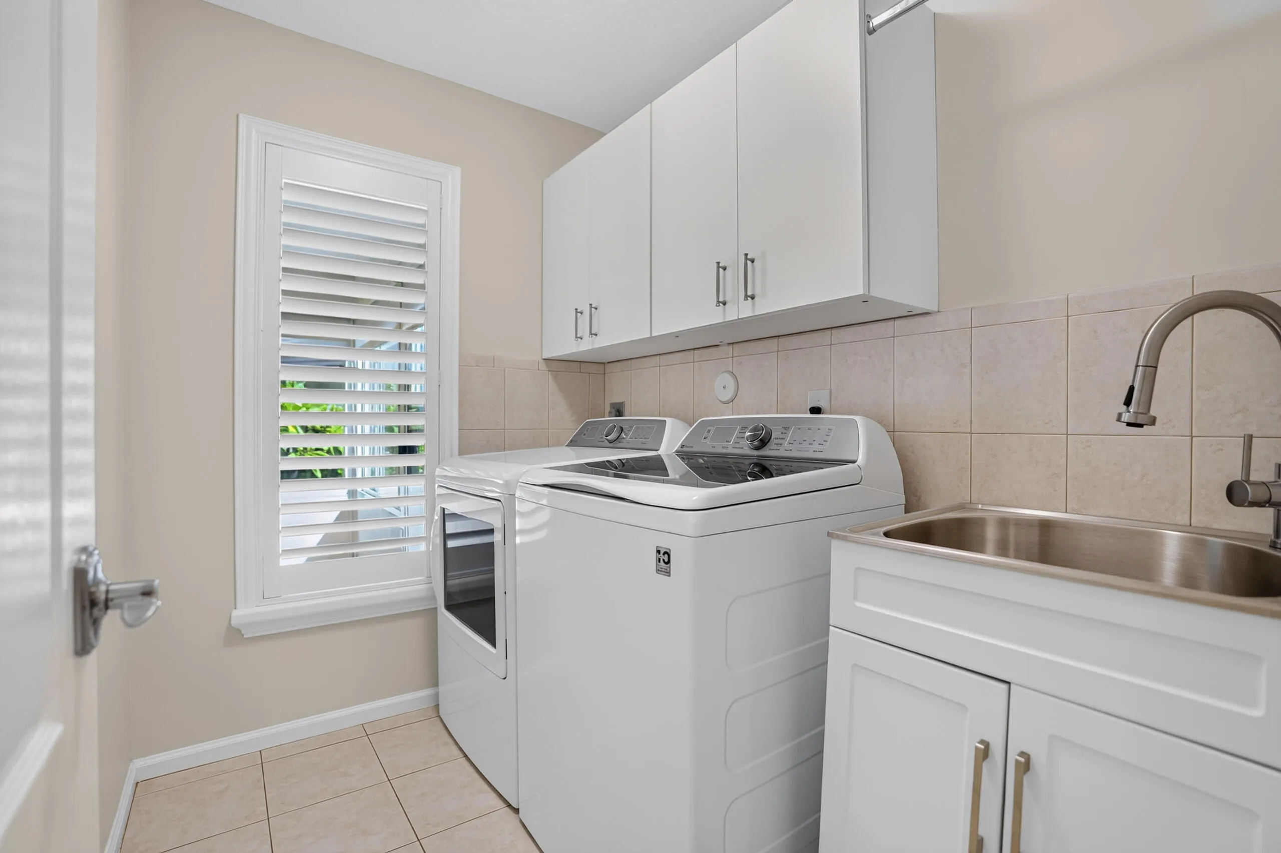 White cabinetry, stainless steel utility sink with a high-arc faucet, tiled flooring, and a window with plantation shutters