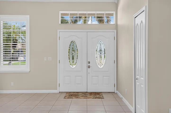 Double entry doors with decorative oval glass inserts, transom window, window with interior shutters, and tile flooring