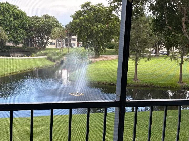 Screened balcony with black railing overlooking a water with fountain, surrounded by manicured lawns and mature trees