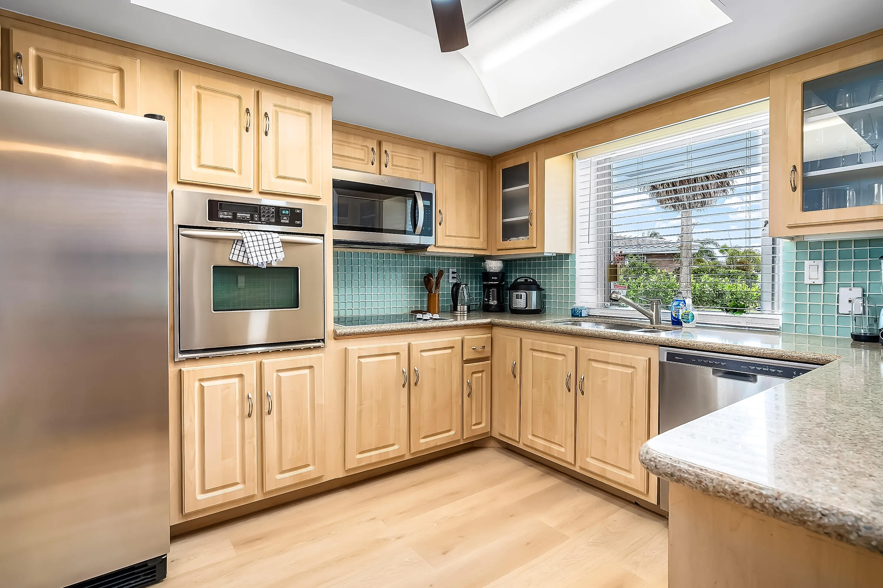 Kitchen featuring light wood-finish cabinetry, stainless steel appliances including a built-in oven and microwave, stone-finish countertops, and a tiled backsplash