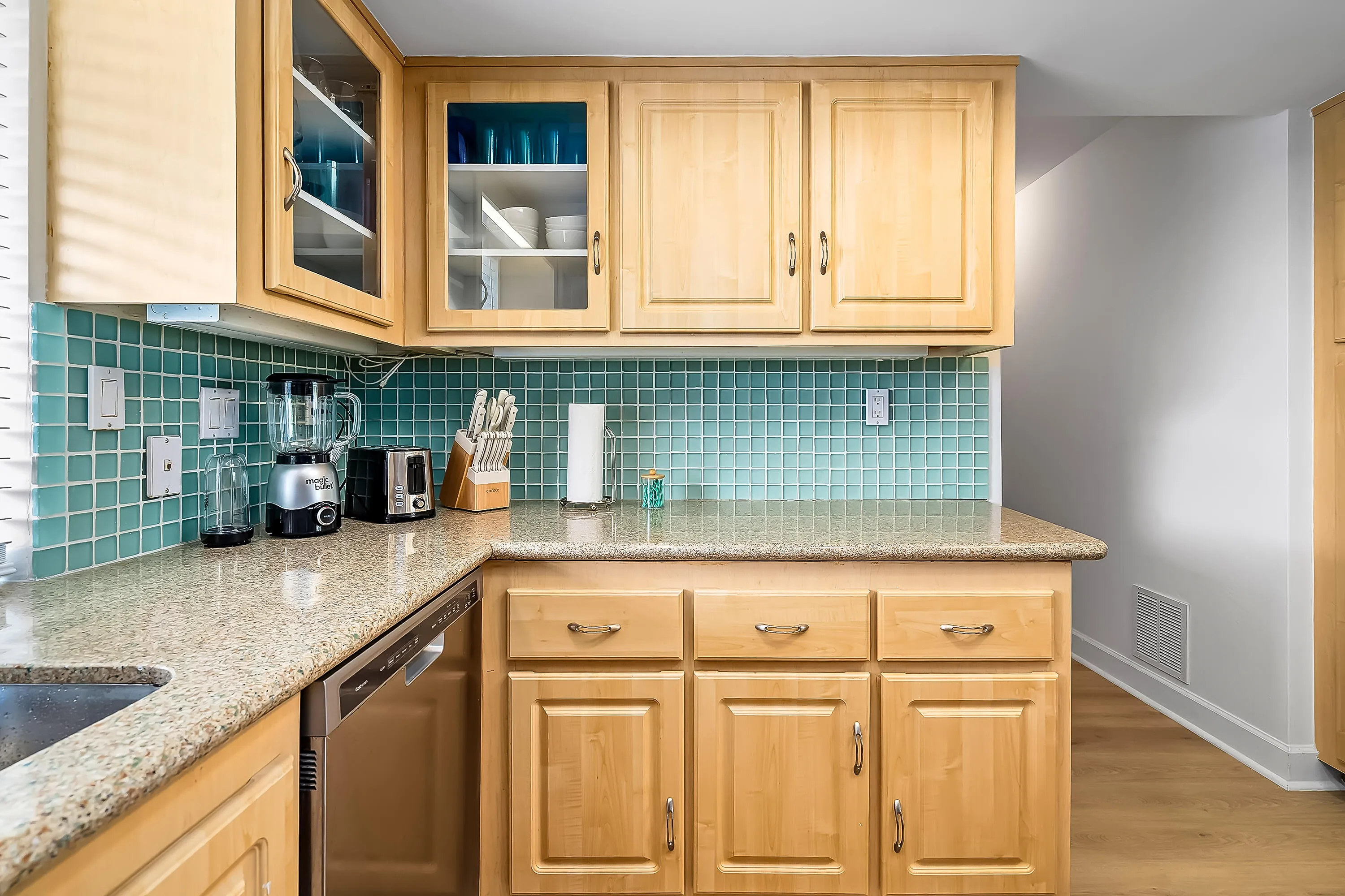 Kitchen featuring light wood cabinetry with glass-front accents, granite-style countertops, a blue-green tile backsplash, and stainless steel dishwasher