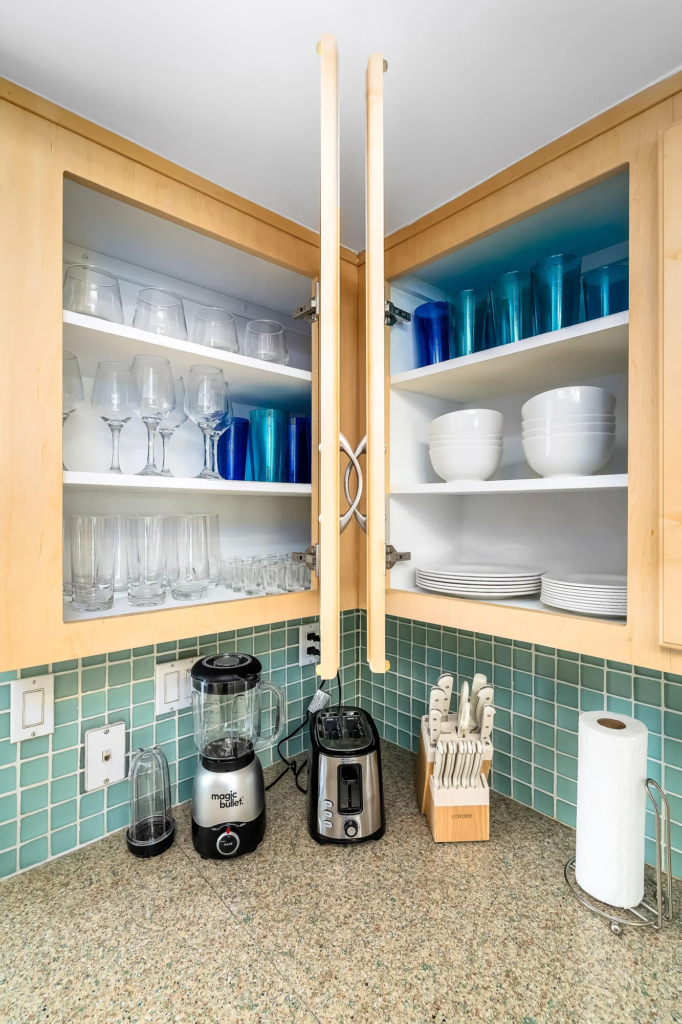 Kitchen countertop featuring a speckled stone-finish surface, light wood-finish cabinetry with open doors, and a turquoise square tile backsplash