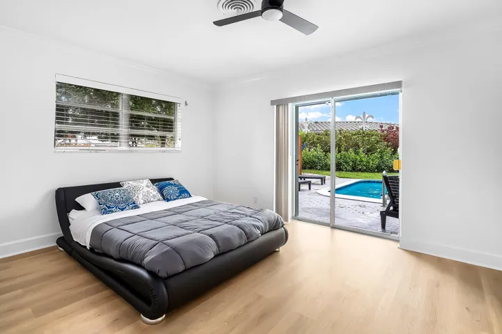 Wood-finish flooring throughout, featuring a ceiling fan, window with horizontal blinds, and a sliding glass door opening to a paved patio and in-ground pool