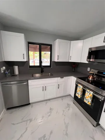 Corner kitchen with white shaker-style cabinetry, gray countertops, stainless steel appliances, and a window above the sink