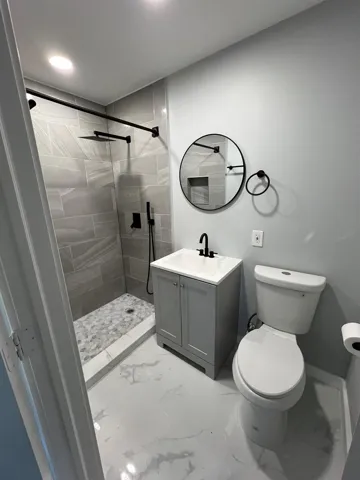 Sleek bathroom featuring a tiled shower with a black rain shower head, a gray vanity with an integrated sink, a round black-framed mirror, and patterned flooring