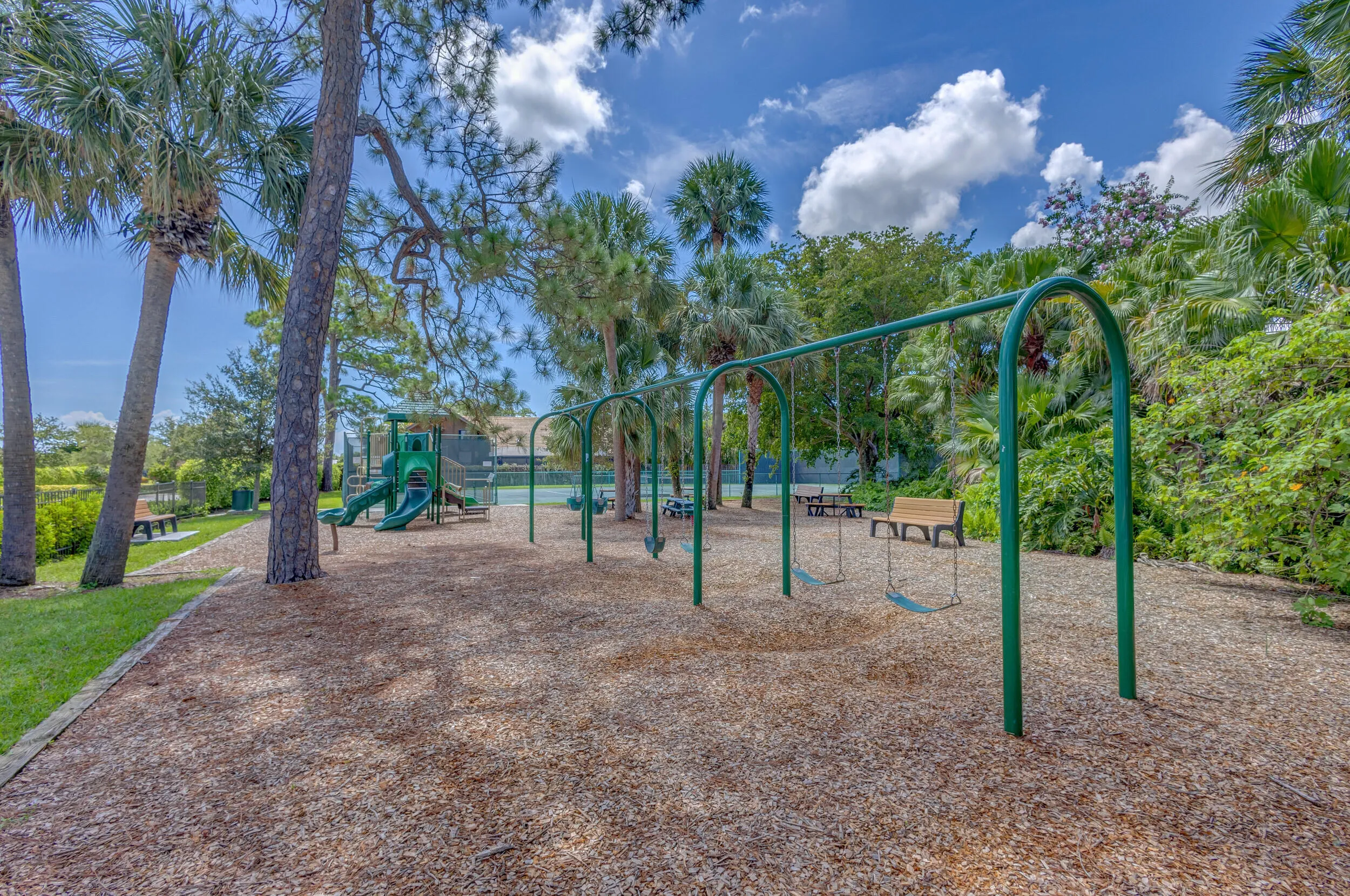 Outdoor playground area with wood chip surfacing, multiple swing sets, a green slide structure, surrounding mature palm trees, and a distant tennis court