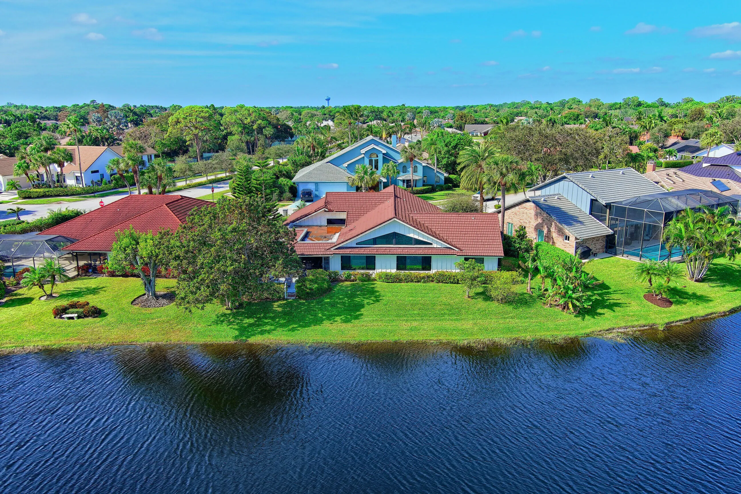 Waterfront property featuring a red tile roof, expansive green lawn, mature landscaping, and screened enclosure