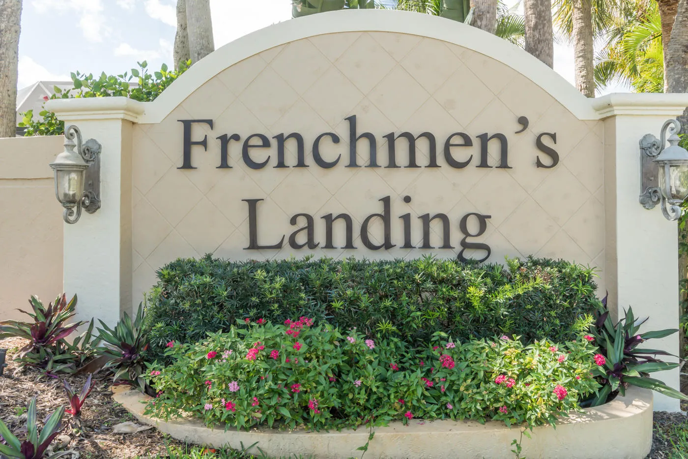 Stone entry monument with sconce lighting, decorative plantings, and tropical landscaping