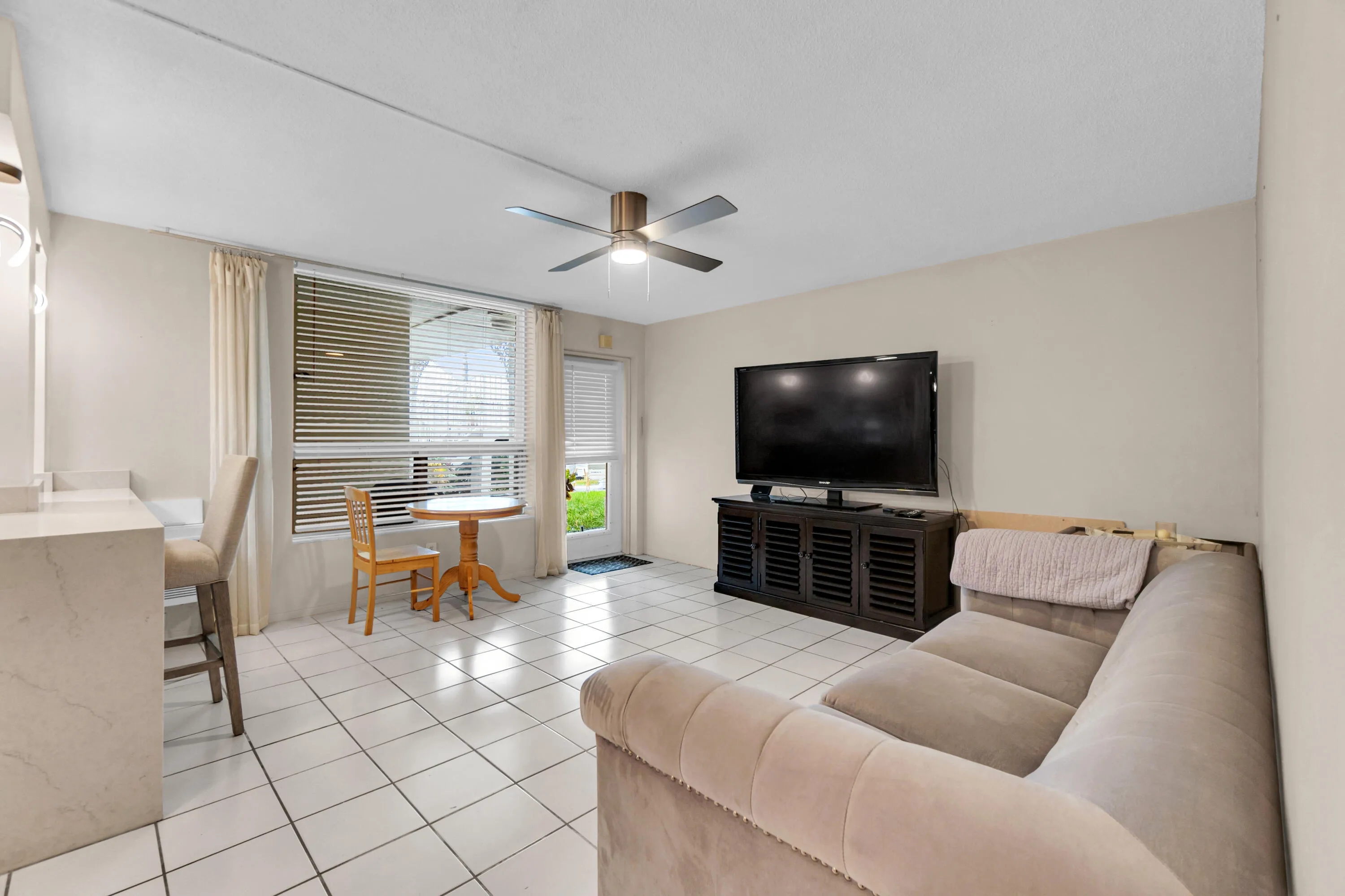 White tile flooring, ceiling fan, built-in breakfast bar with stone-finish countertop, large window with blinds, and glass-paneled exterior door