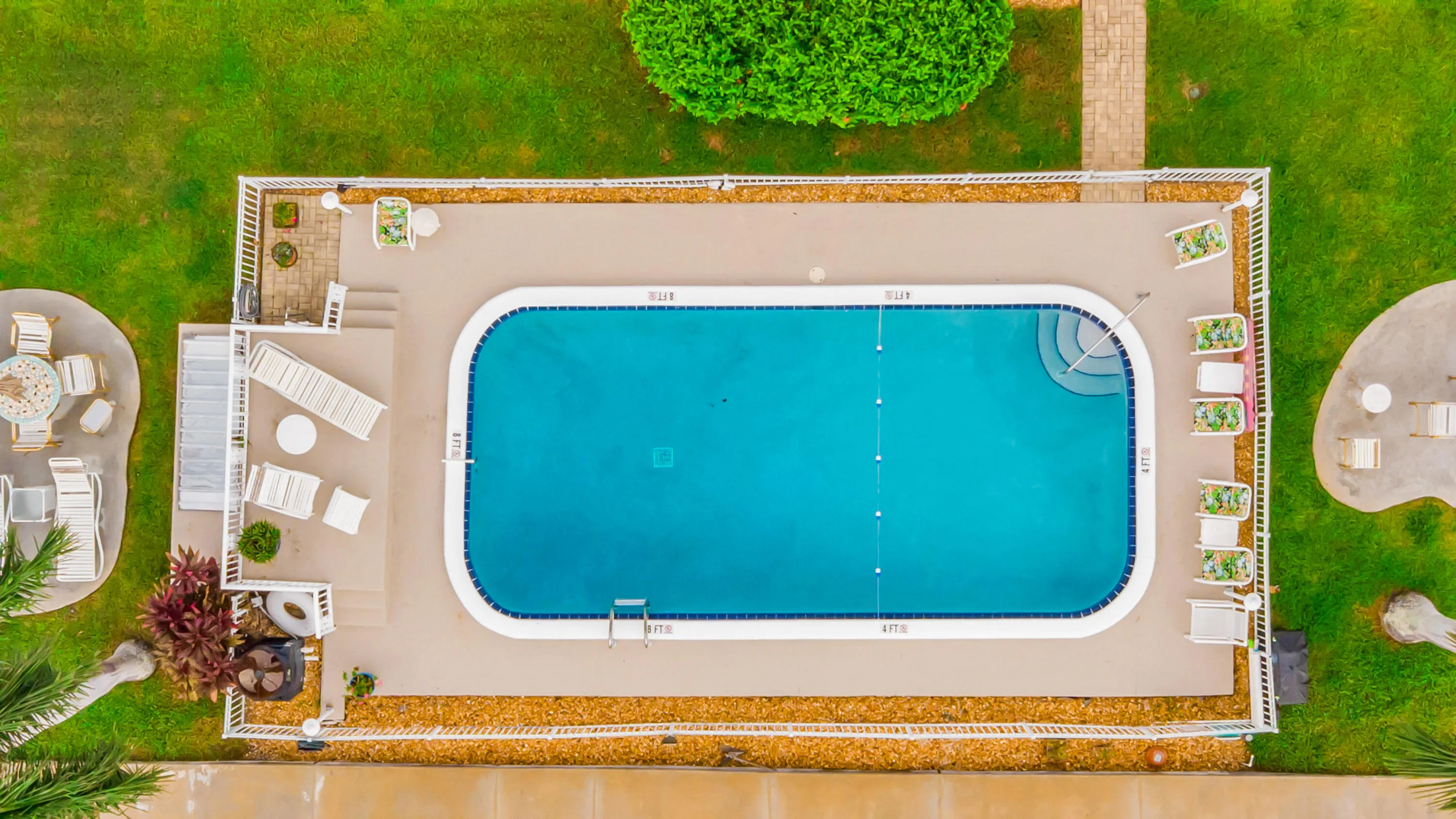 Rectangular swimming pool with integrated steps, surrounded by a paved deck, white fencing, and landscaped greenery