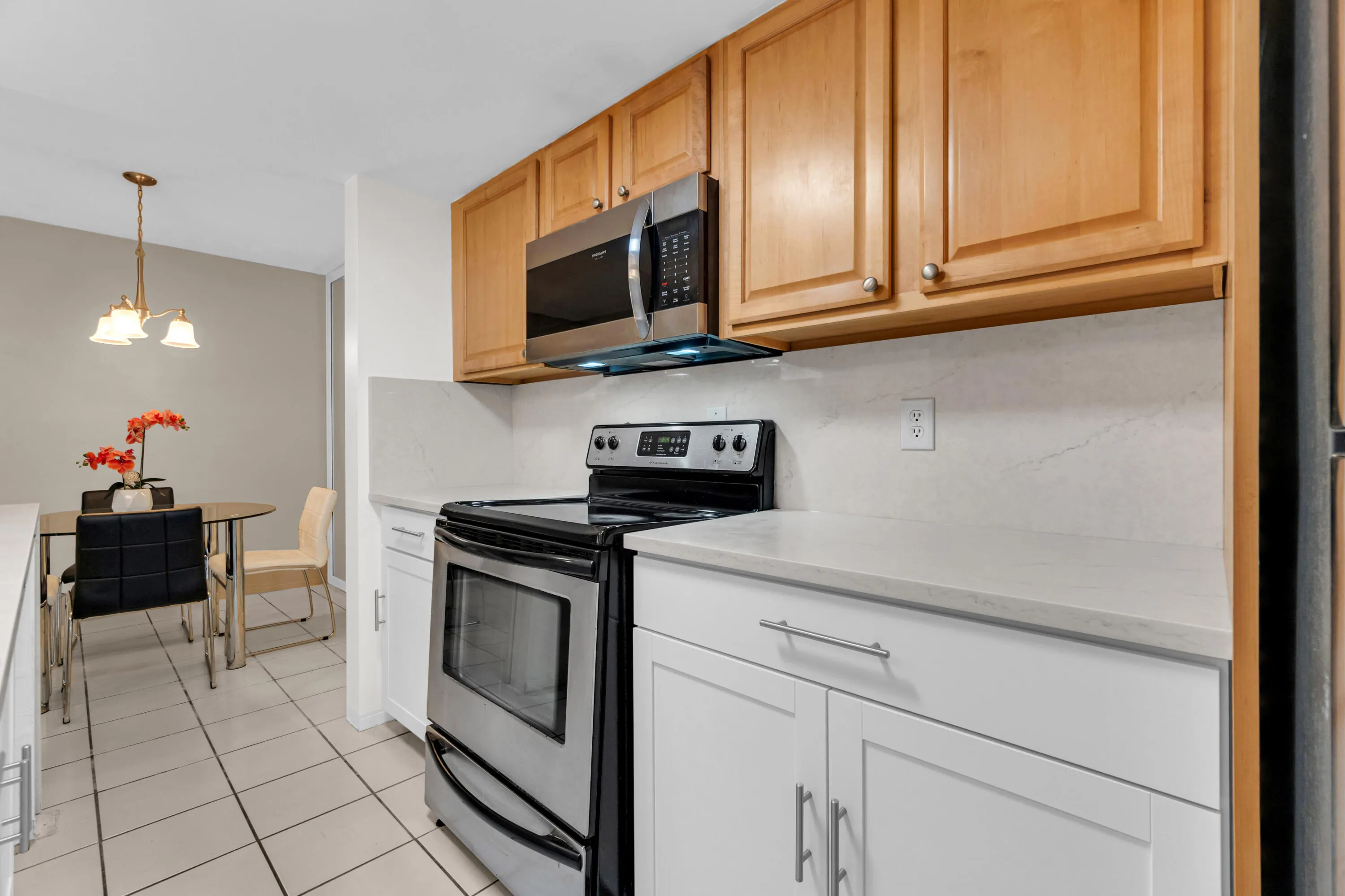 Kitchen featuring stainless steel appliances, light wood-finish upper cabinetry, white lower cabinetry, and light-toned countertops with a matching backsplash