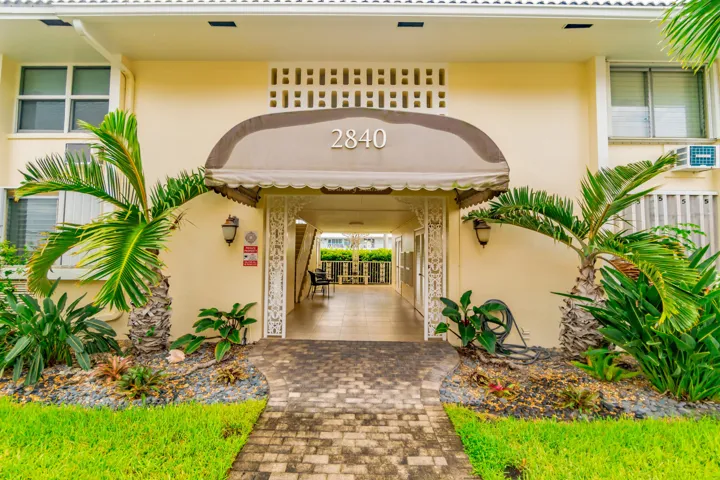 Building exterior with a covered entry, featuring a paved walkway, manicured lawn, and tropical landscaping