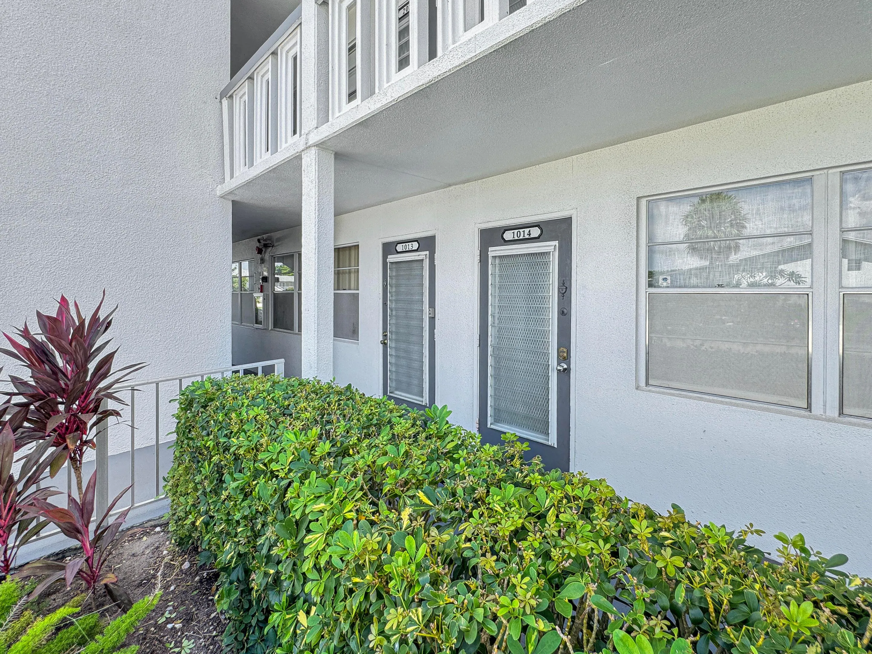 Building exterior with a textured finish, balcony with white railings, and manicured landscaping featuring various shrubs and plants