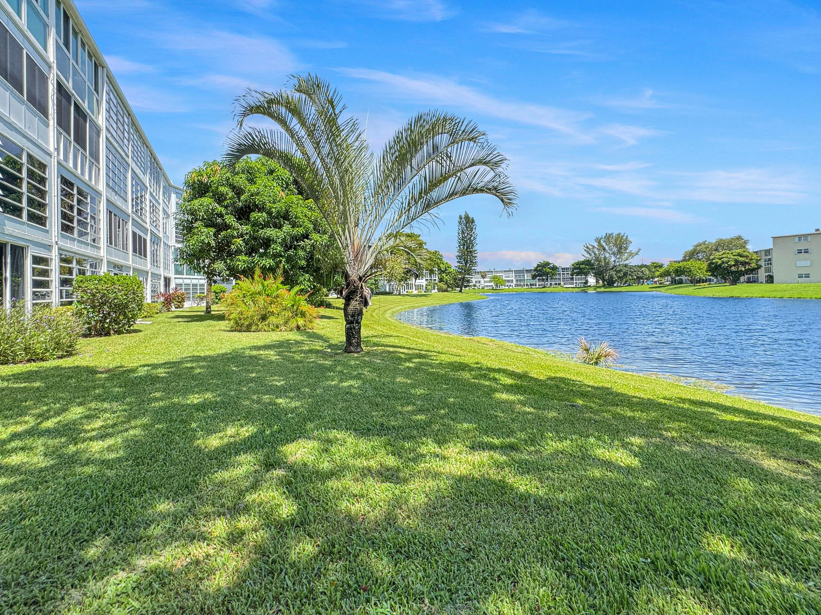 Lakeside building with multiple windows, surrounded by green lawns, mature palm trees, and established landscaping