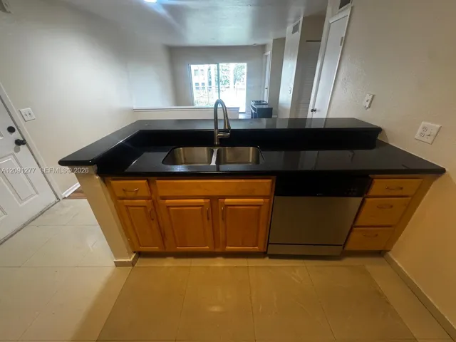 Kitchen peninsula featuring black countertops, a stainless steel dual-basin undermount sink with a high-arch faucet, wood-finish cabinetry, and a built-in stainless steel dishwasher