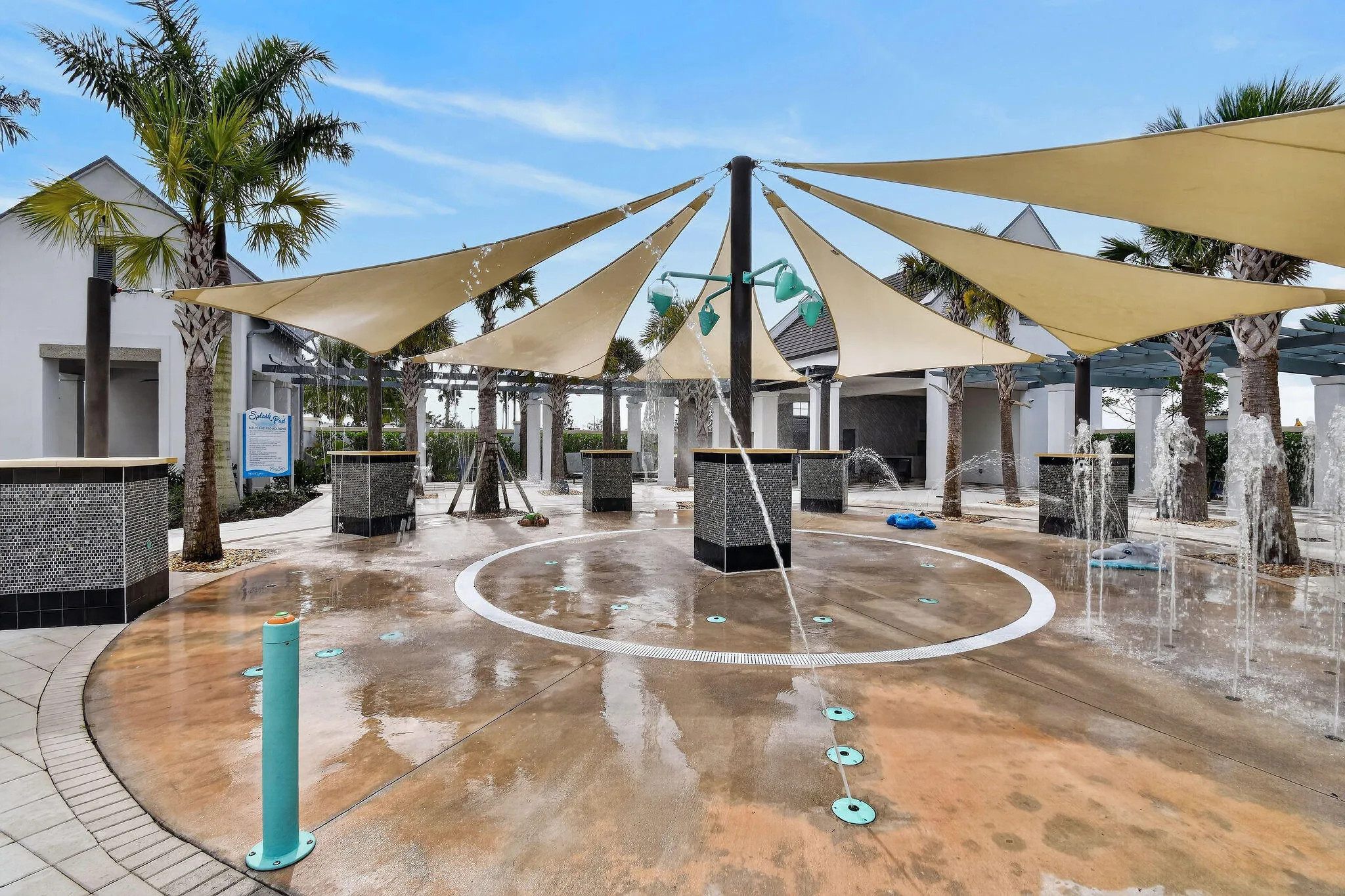 Splash pad with multiple water features, shade sails, surrounding palm trees, and tiled accent pillars