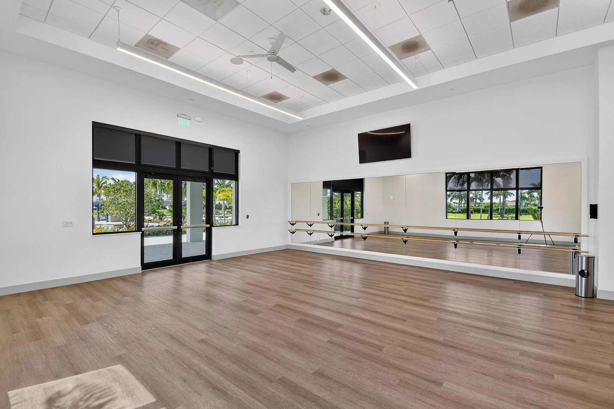Spacious room featuring wood-finish flooring, full-wall mirrors with ballet barres, a ceiling fan, and large windows with exterior access