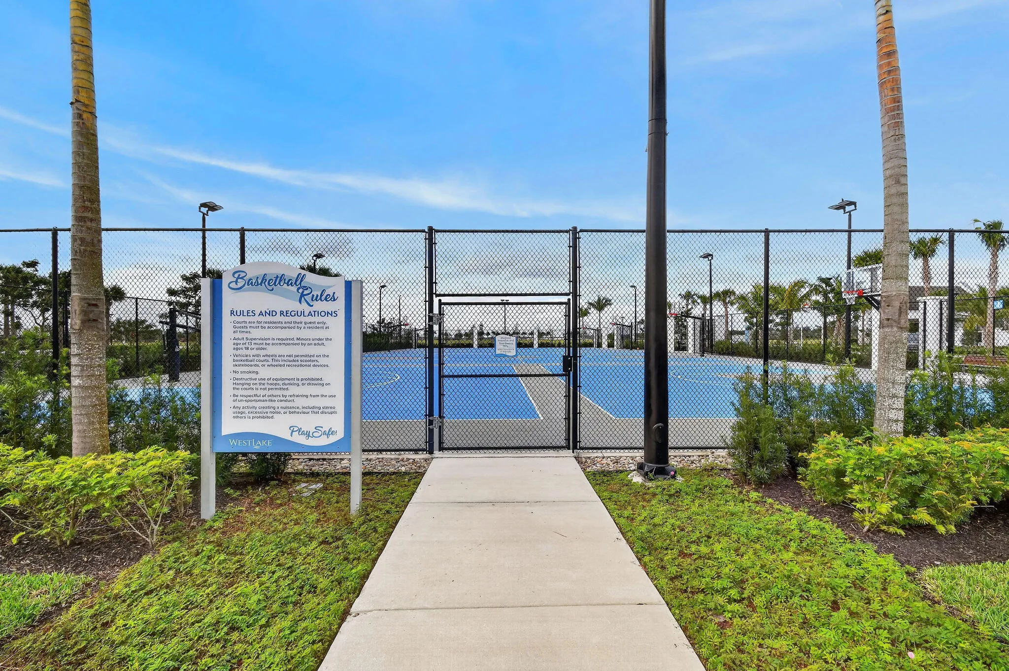 Outdoor basketball court with a chain-link fence enclosure, paved walkway, and landscaping