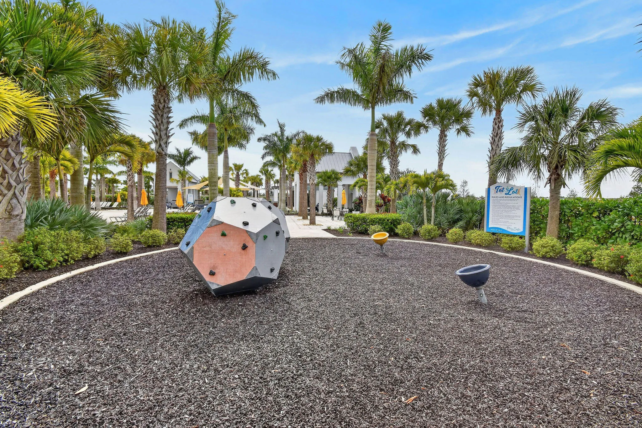 Outdoor play area with rubberized surfacing, climbing structure, and sound tubes, surrounded by palm trees and landscaping