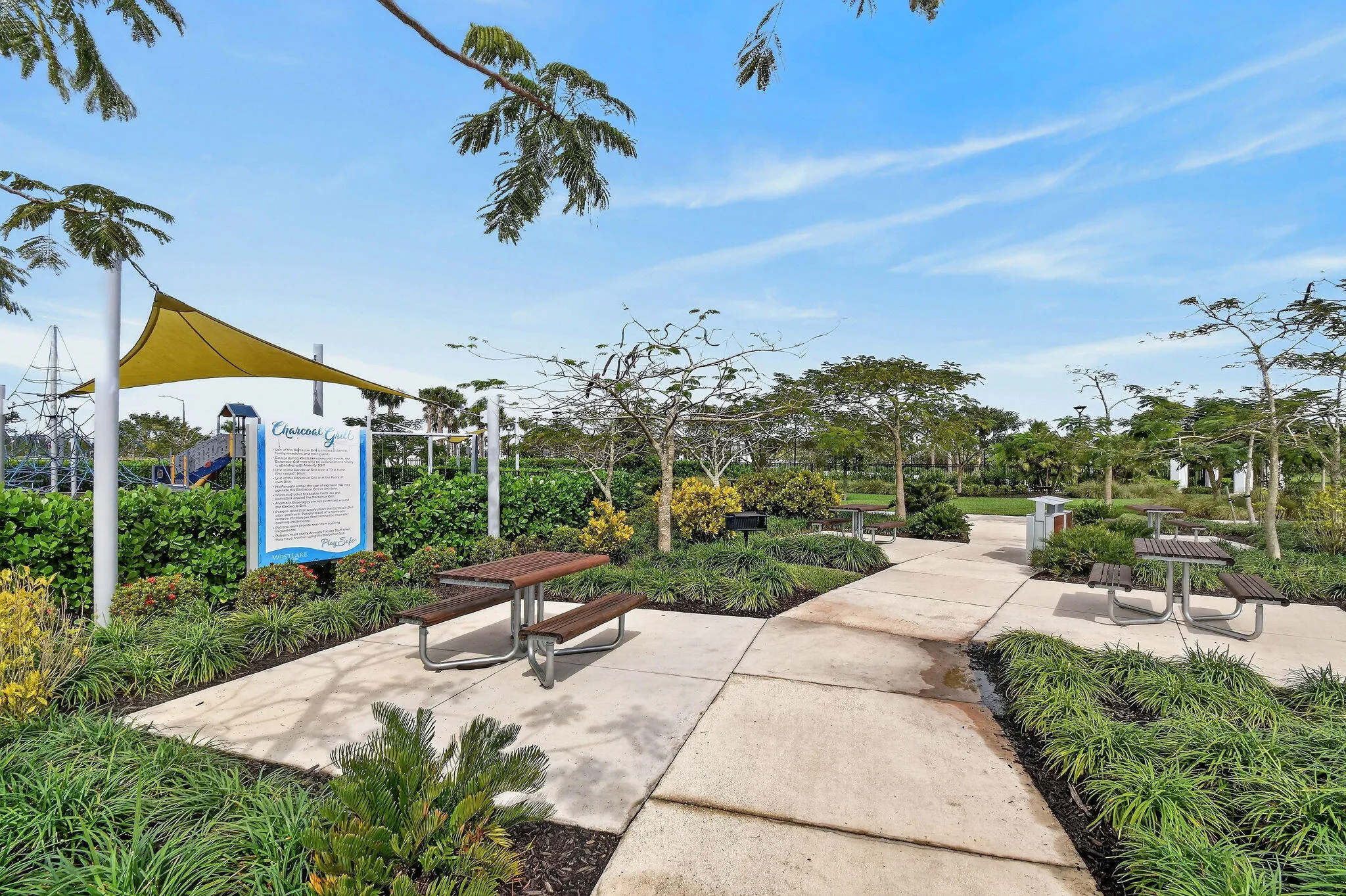 Paved pathways traversing a landscaped park, featuring multiple picnic tables, a shade structure, and a prominent informational sign