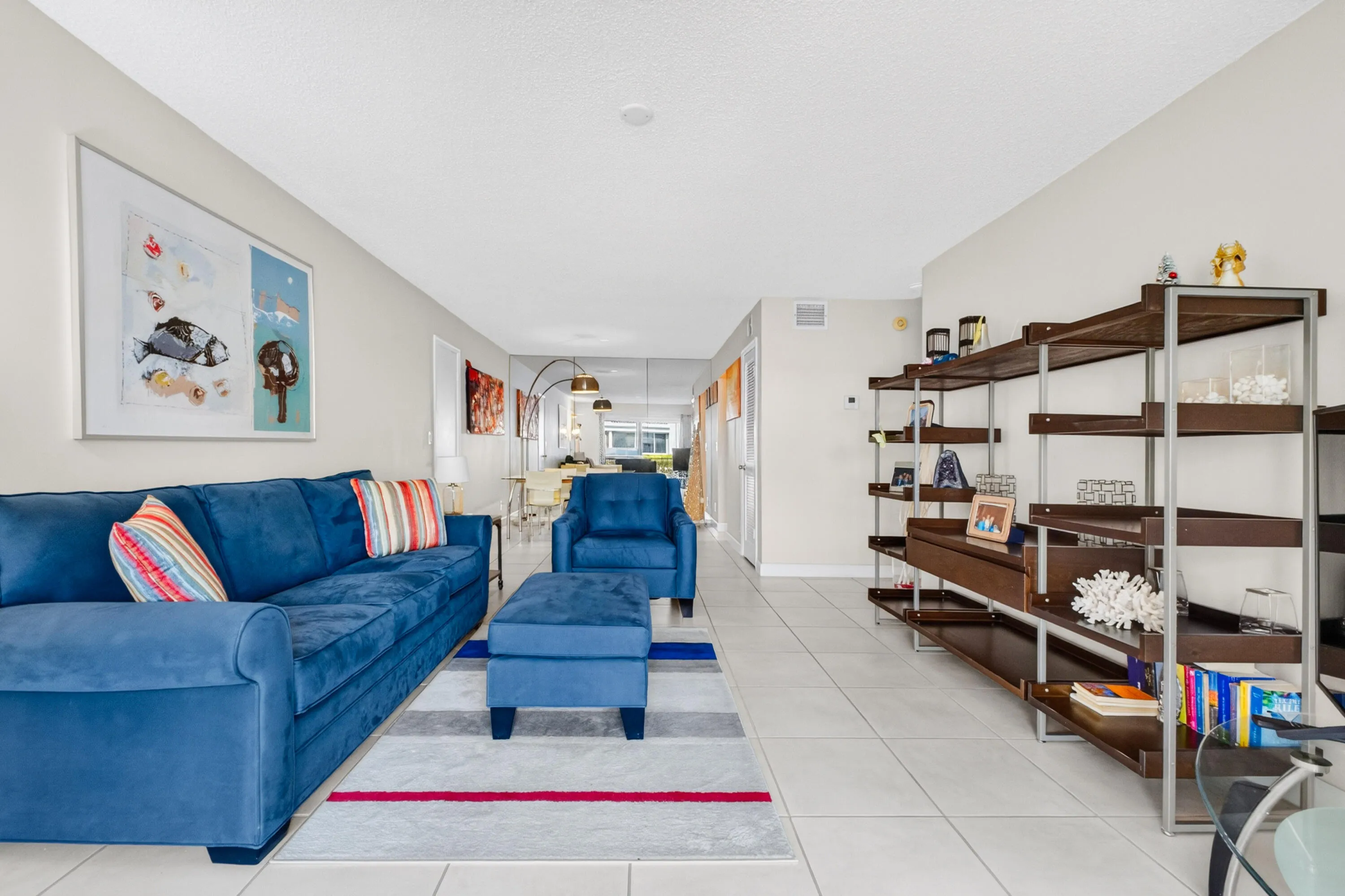 Bright, open-concept living area featuring tile flooring throughout, custom wood-finish shelving with ample display and storage capacity, and a vibrant blue upholstered sofa anchoring the space