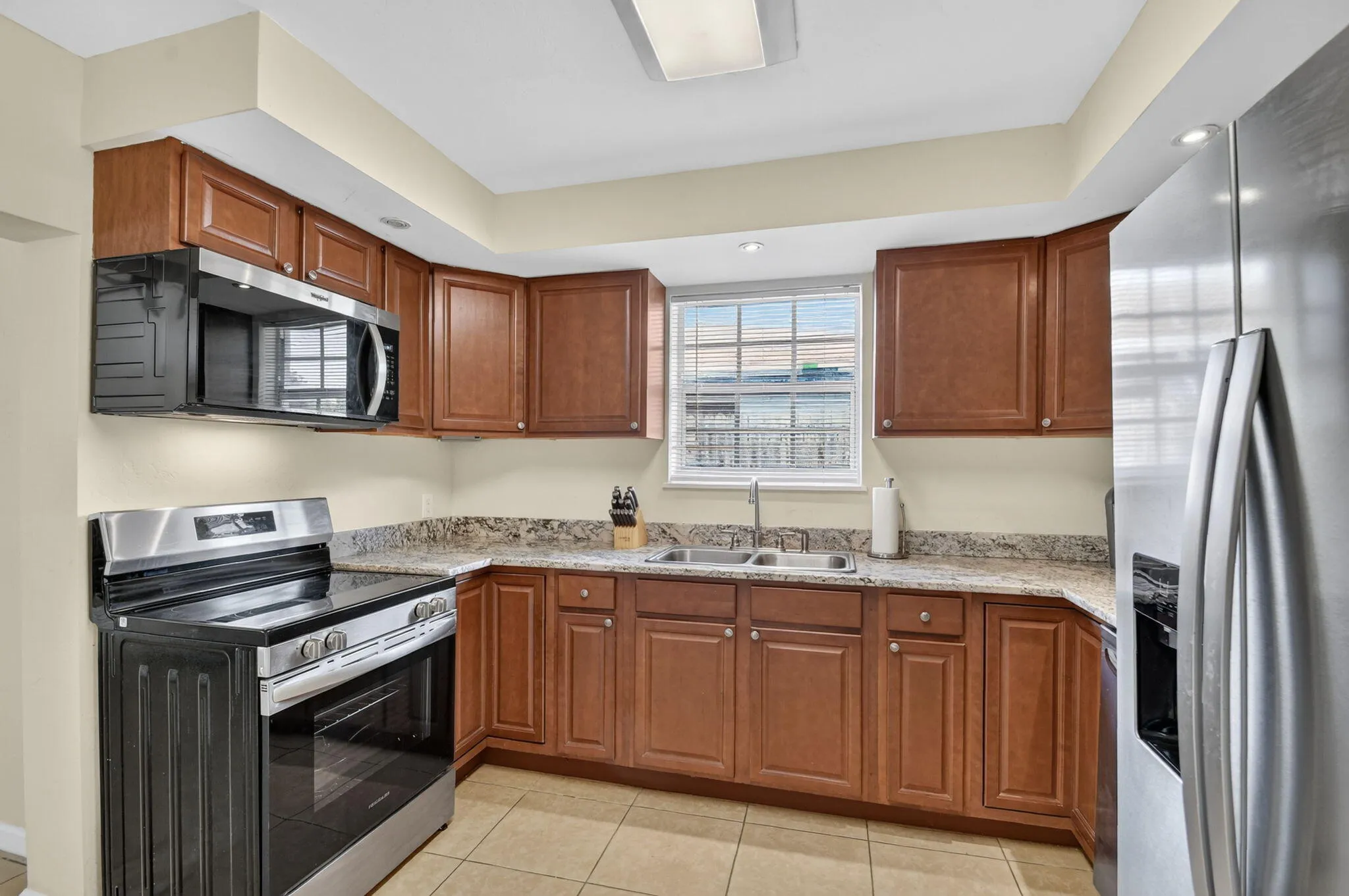 Kitchen featuring wood-finish cabinetry, stone-finish countertops, stainless steel appliances, a double basin sink, and tiled flooring